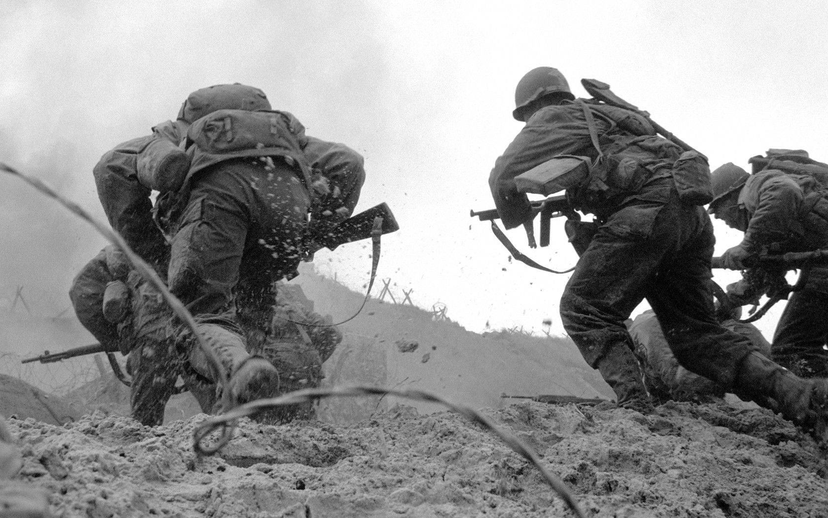 Ww2 桌布 壁紙 — 身穿重型裝備的士兵在泥濘地形中衝鋒，鐵絲網和泥土在混亂的戰場場景中飛揚。