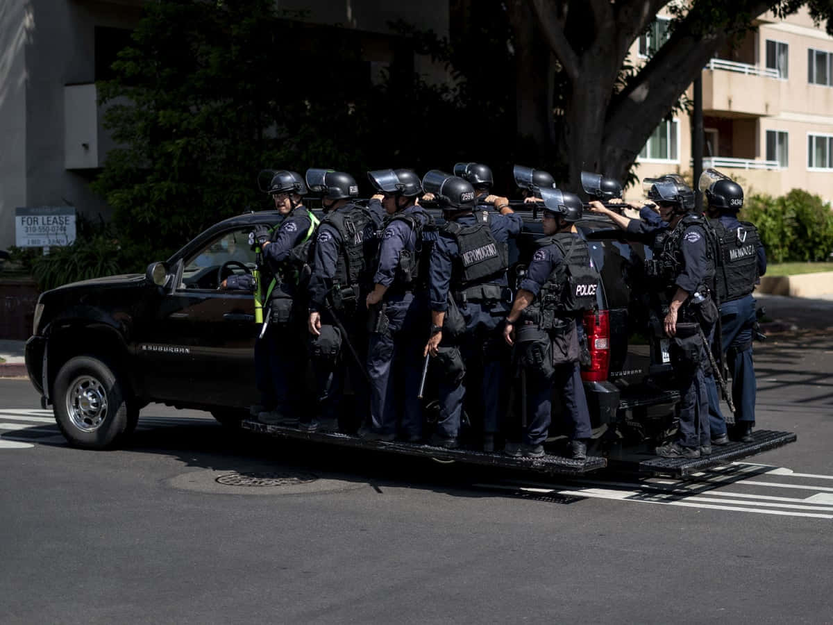 警察 桌布 壁紙 — 身穿防暴裝備的警察在陽光明媚的城市街道上，在一輛黑色皮卡車的後方列隊。