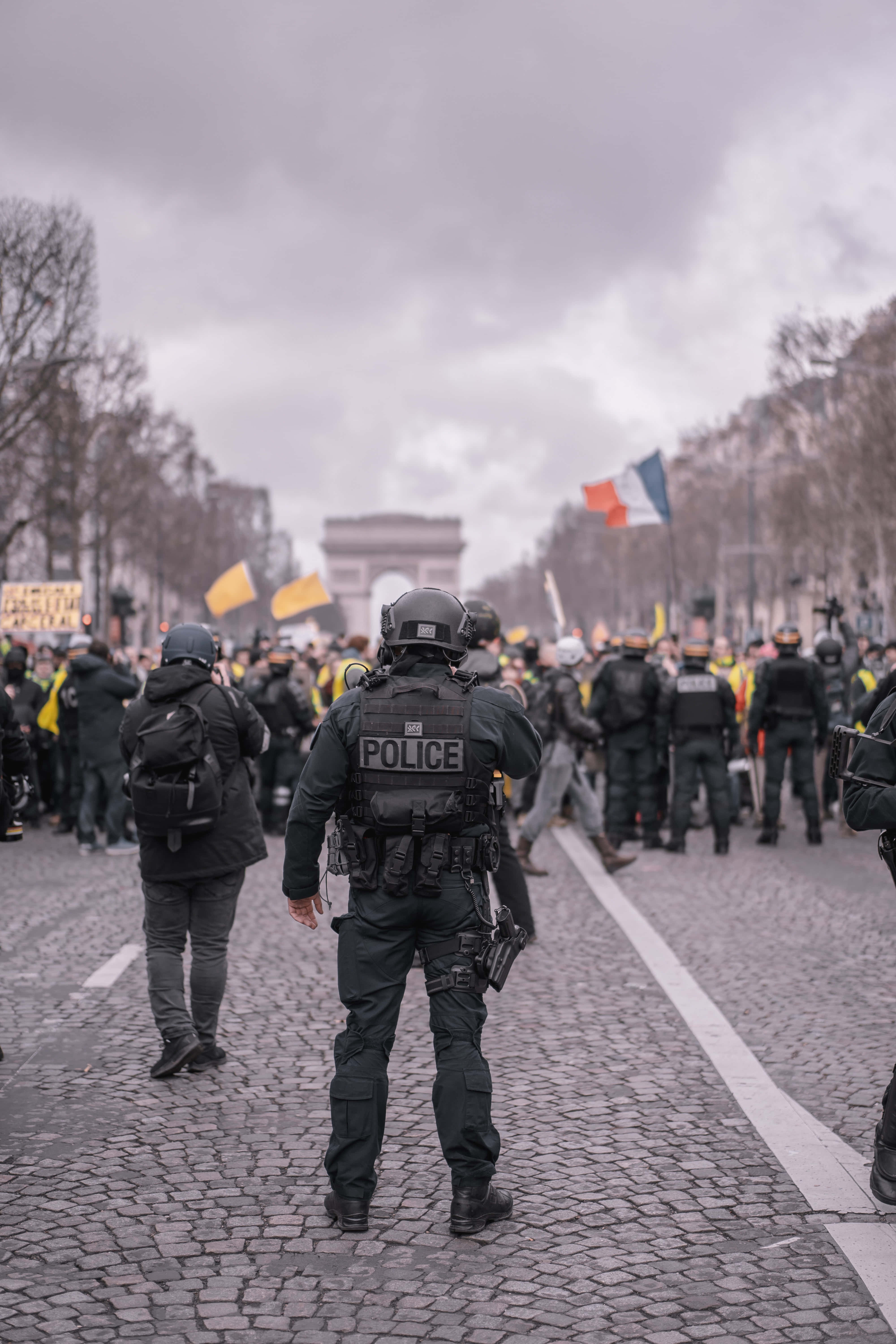 Law Enforcement 桌布 壁紙 — 身穿防暴裝備的警察站在鵝卵石街道上，面對著一群舉著黃色旗幟的人群，天空多雲，靠近凱旋門。