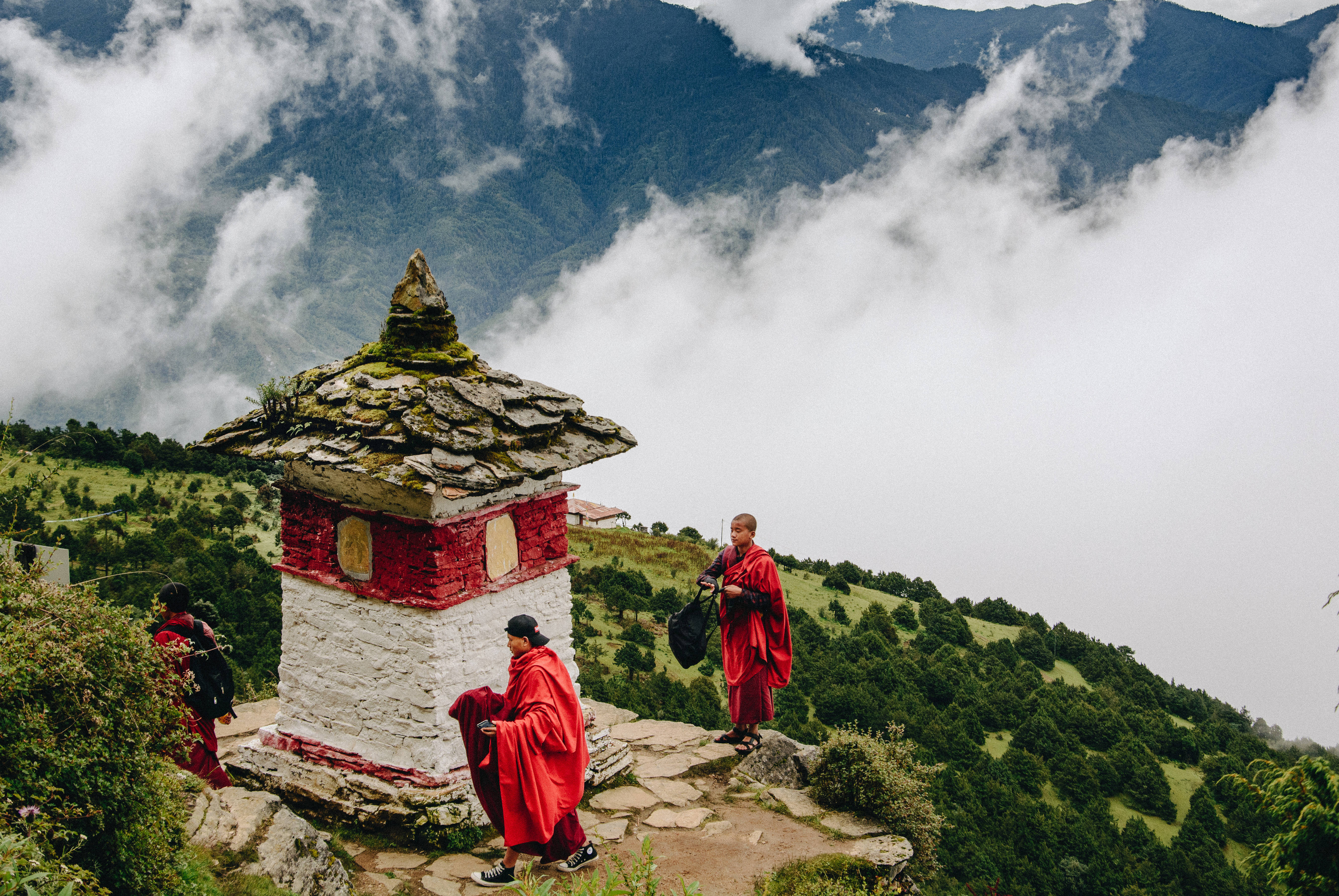 Bhutan 桌布 壁紙 — 身穿鮮紅色僧袍的僧侶在雲霧繚繞的山脊上，沿著一座石砌佛塔行走，俯瞰雲海覆蓋的山谷。