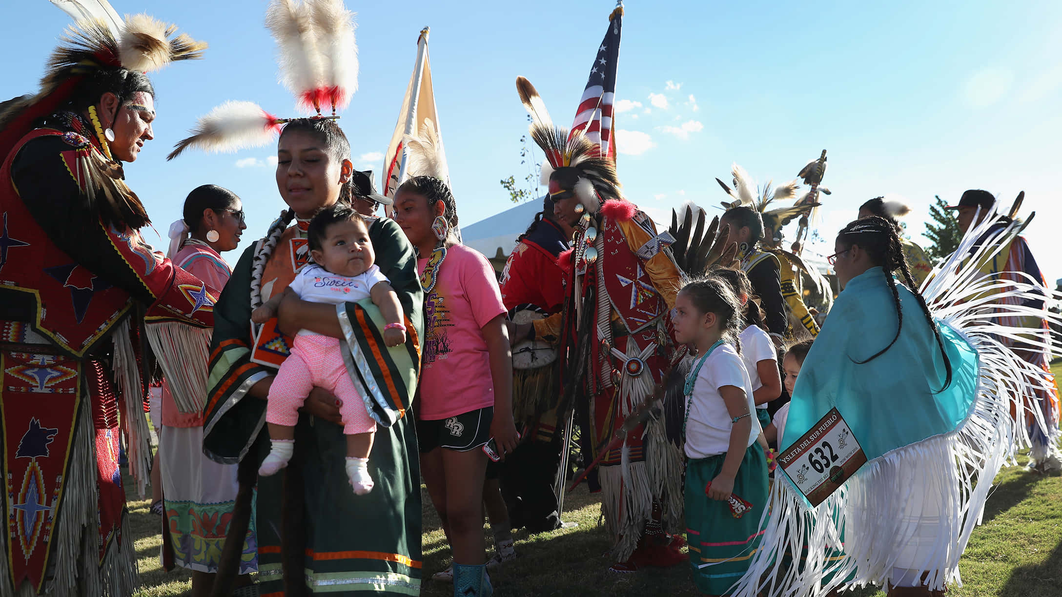 Native American 桌布 壁紙 — 身穿鮮豔傳統服飾、頭戴羽毛頭飾的美洲原住民舞者，在晴朗藍天下於戶外文化儀式中聚集。