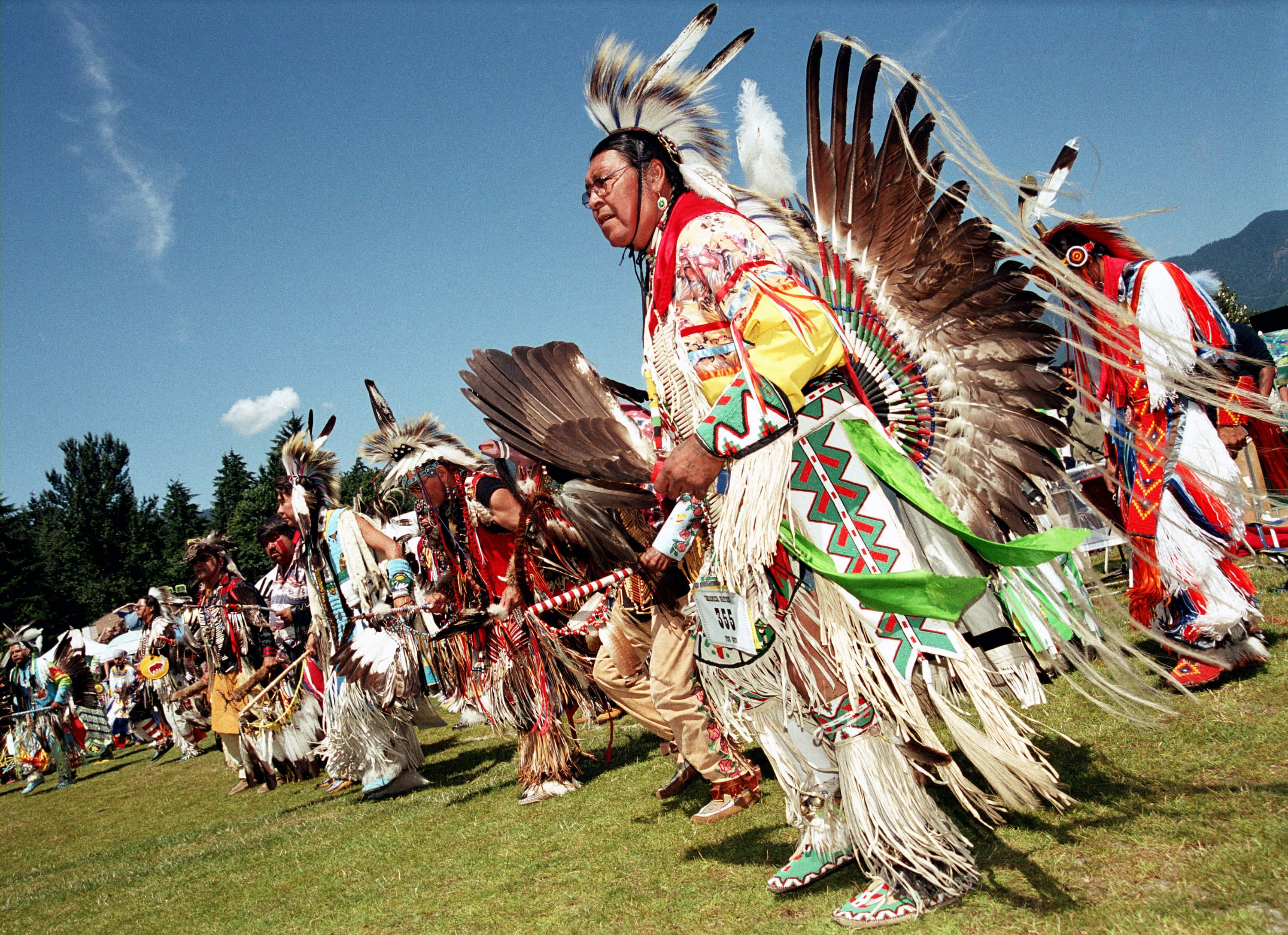 American Indian 桌布 壁紙 — 身穿鮮豔服飾、頭戴羽毛頭飾的美洲原住民舞者在晴朗的藍天下進行傳統儀式。