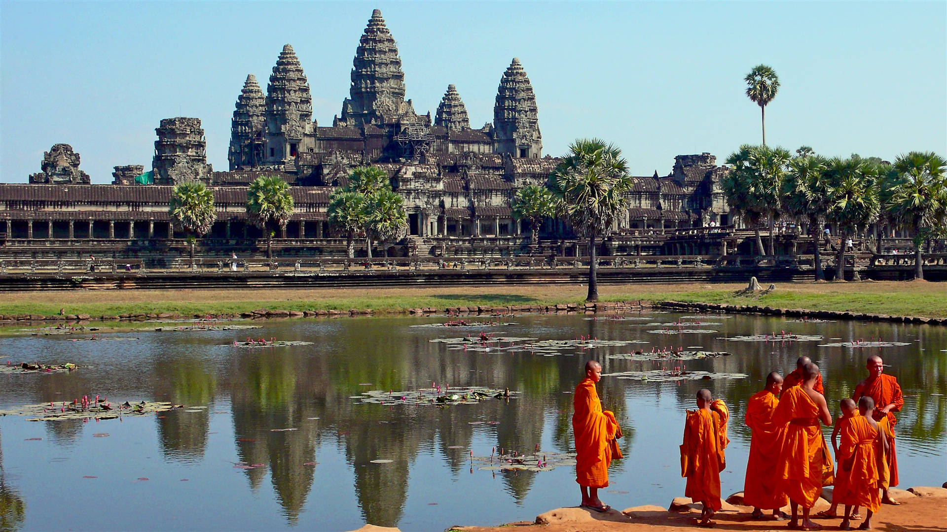 Cambodia 桌布 壁紙 — 身穿鮮豔橙色僧袍的僧侶沿著池塘邊行走，雄偉的吳哥窟寺廟群在清澈藍天下倒映在平靜的水面上。