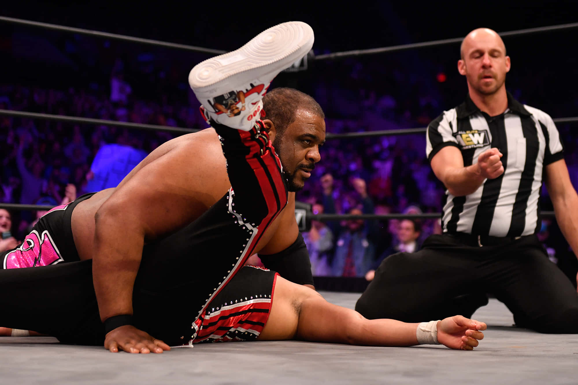 WWE 桌布 壁紙 — 身穿黑色和紅色裝備的摔角手在墊子上執行一個戲劇性的動作，裁判在擠滿人的場館中專注地觀看。