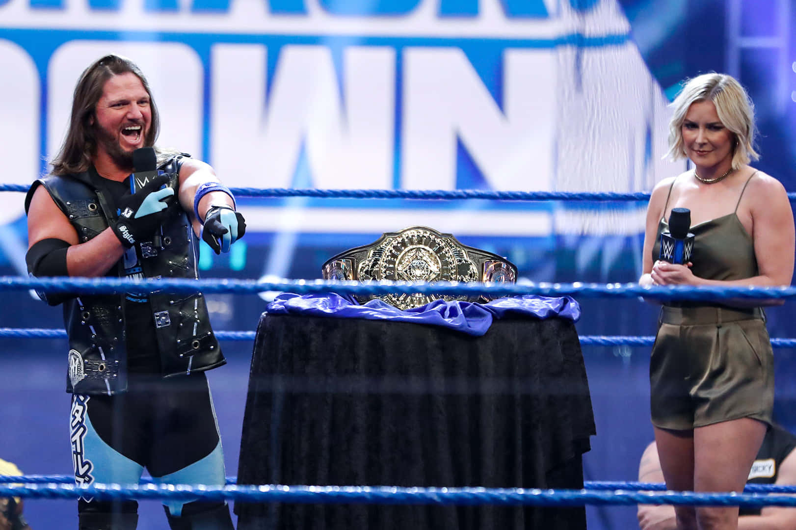 WWE 桌布 壁紙 — 身穿黑色和藍色裝備的摔角手在女主持人旁邊勝利地咆哮，冠軍腰帶放在擂台上的桌子上。