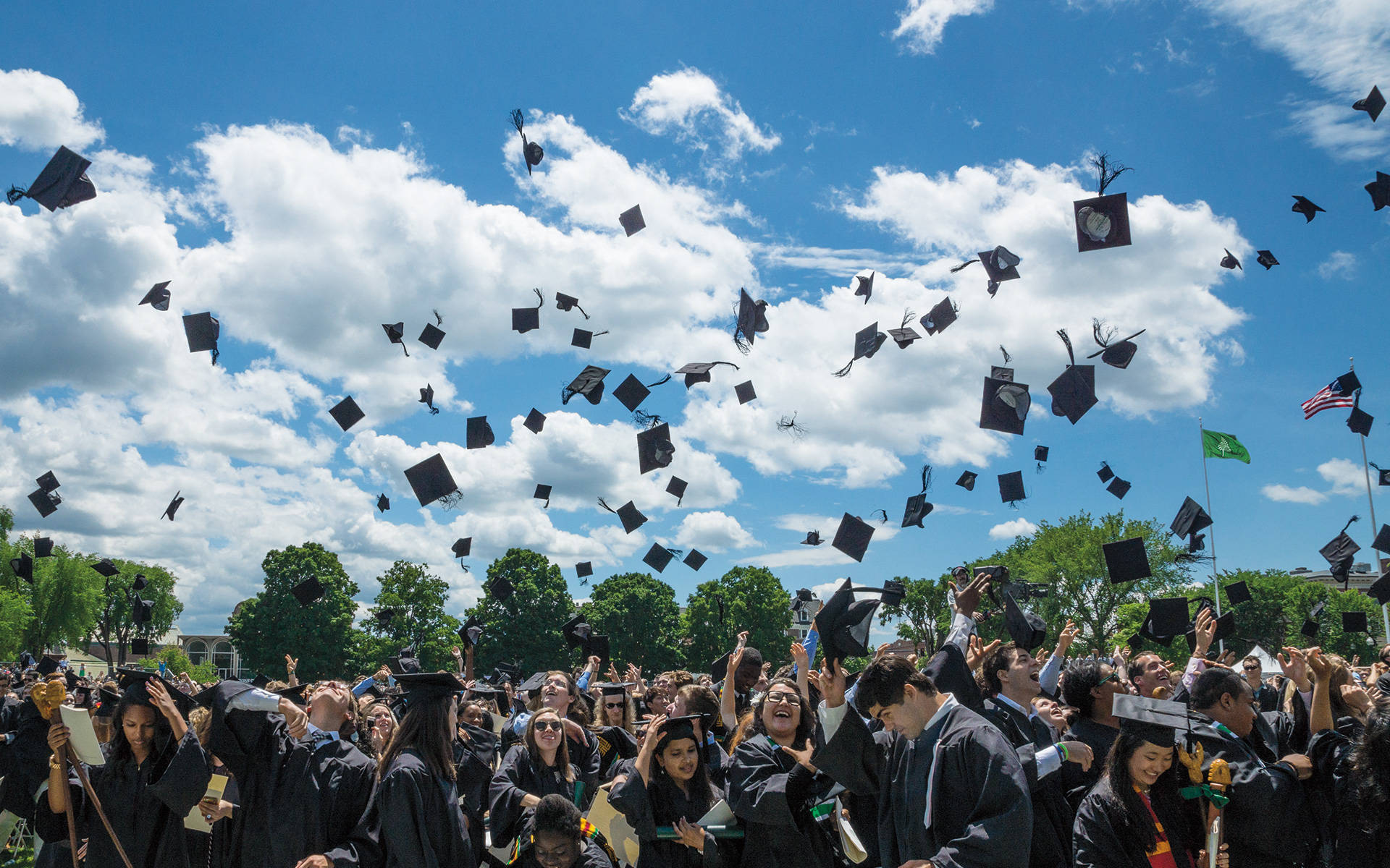 Graduation 桌布 壁紙 — 身穿黑色學士帽和畢業袍的畢業生，歡快地將四方帽拋向點綴著蓬鬆白雲的明亮藍天。