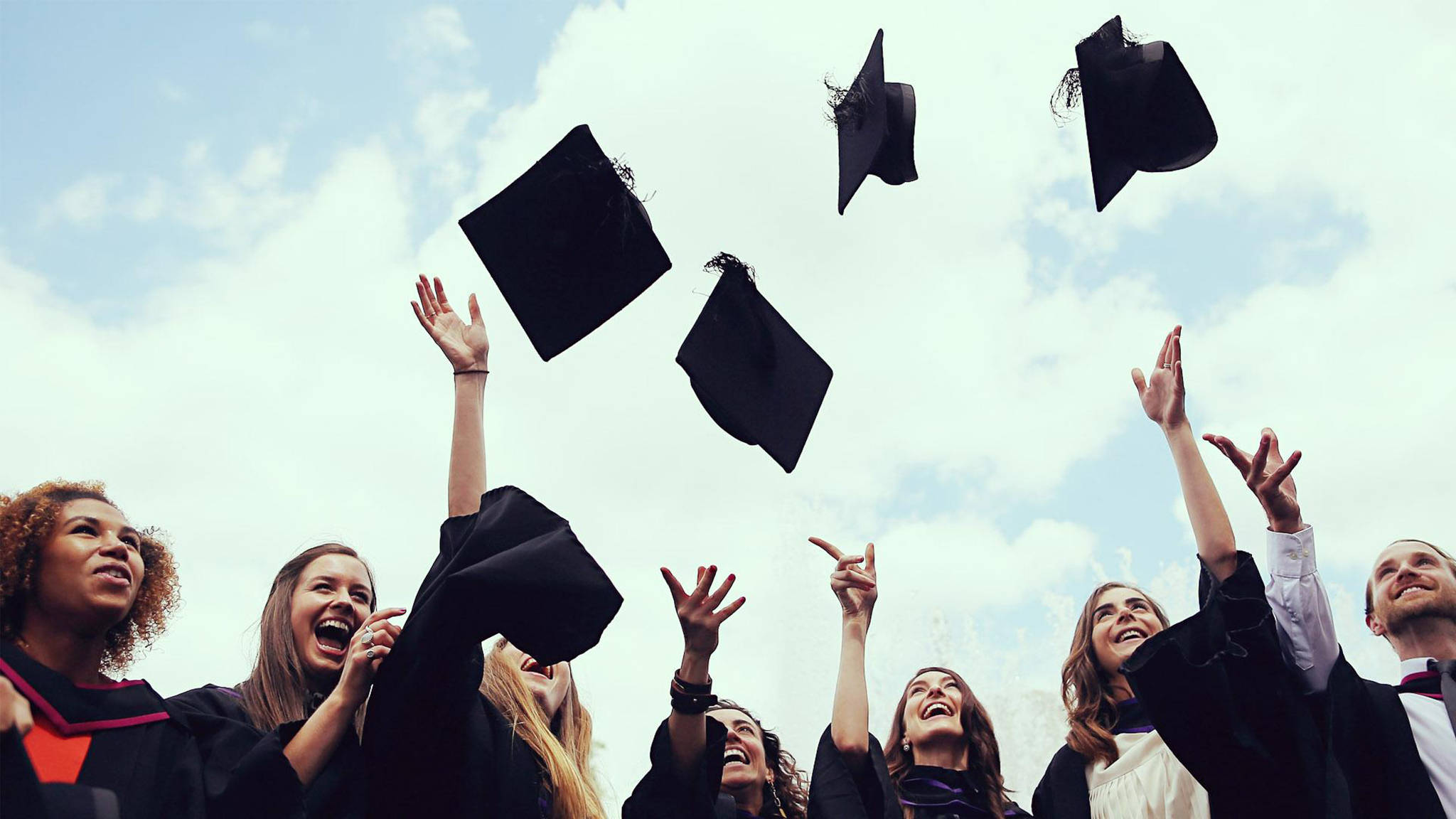 Graduation 桌布 壁紙 — 身穿黑色學士帽和畢業袍的畢業生，在晴朗多雲的天空下，歡快地將四方帽拋向天空。