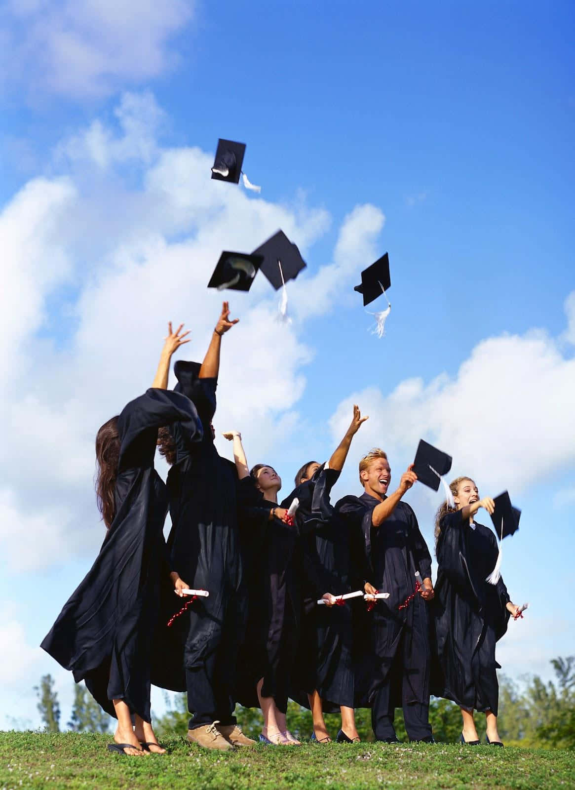 Graduation 桌布 壁紙 — 身穿黑色學士袍和方帽的畢業生，在晴朗的日子裡歡快地將方帽拋向明亮的藍天。