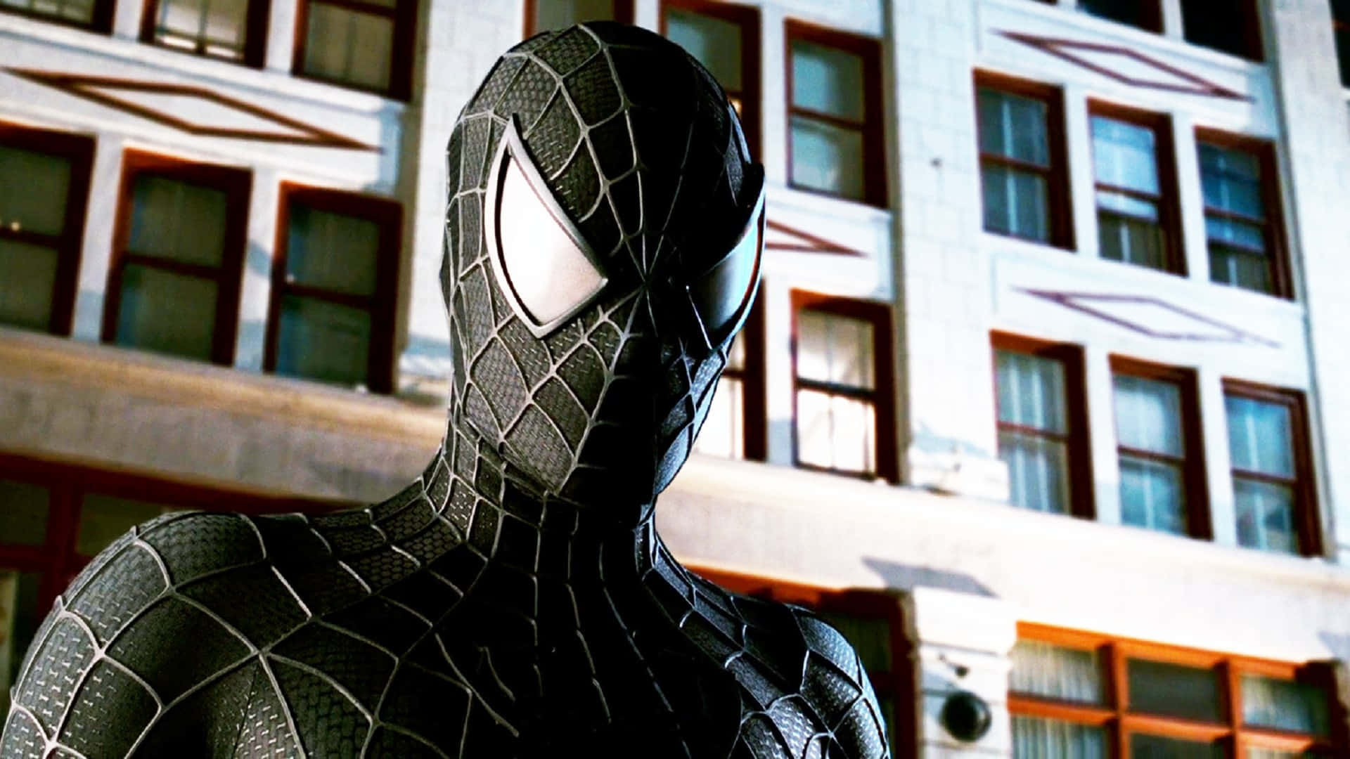 Spider Man 3 桌布 壁紙 — 身穿黑色戰衣的蜘蛛人站在陽光普照的城市建築前，戰衣上有精細的網紋圖案，眼睛發出白光。
