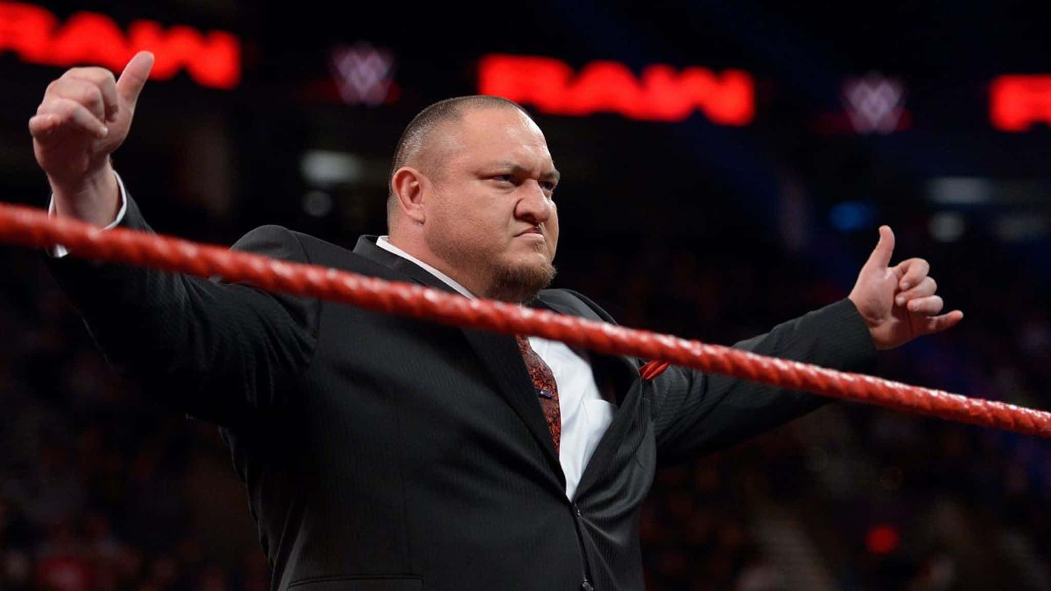 WWE 桌布 壁紙 — 身穿黑色西裝的摔角手在紅色擂台內豎起兩個大拇指，身後有「RAW」燈光閃耀。