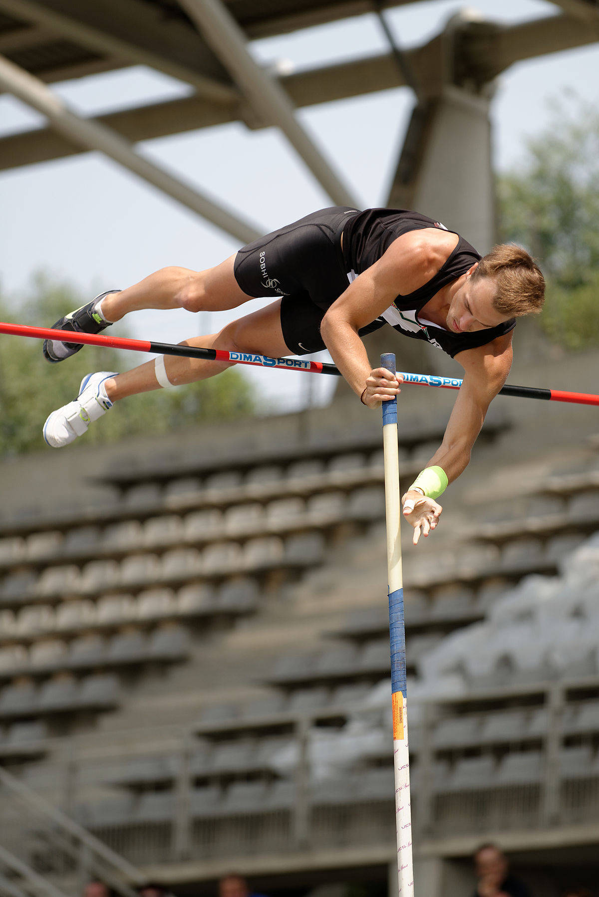 Athletic 桌布 壁紙 — 身穿黑色運動服的運動員在明亮的日光下，以撐竿跳竿越過跳高橫桿，背景是體育場看台。