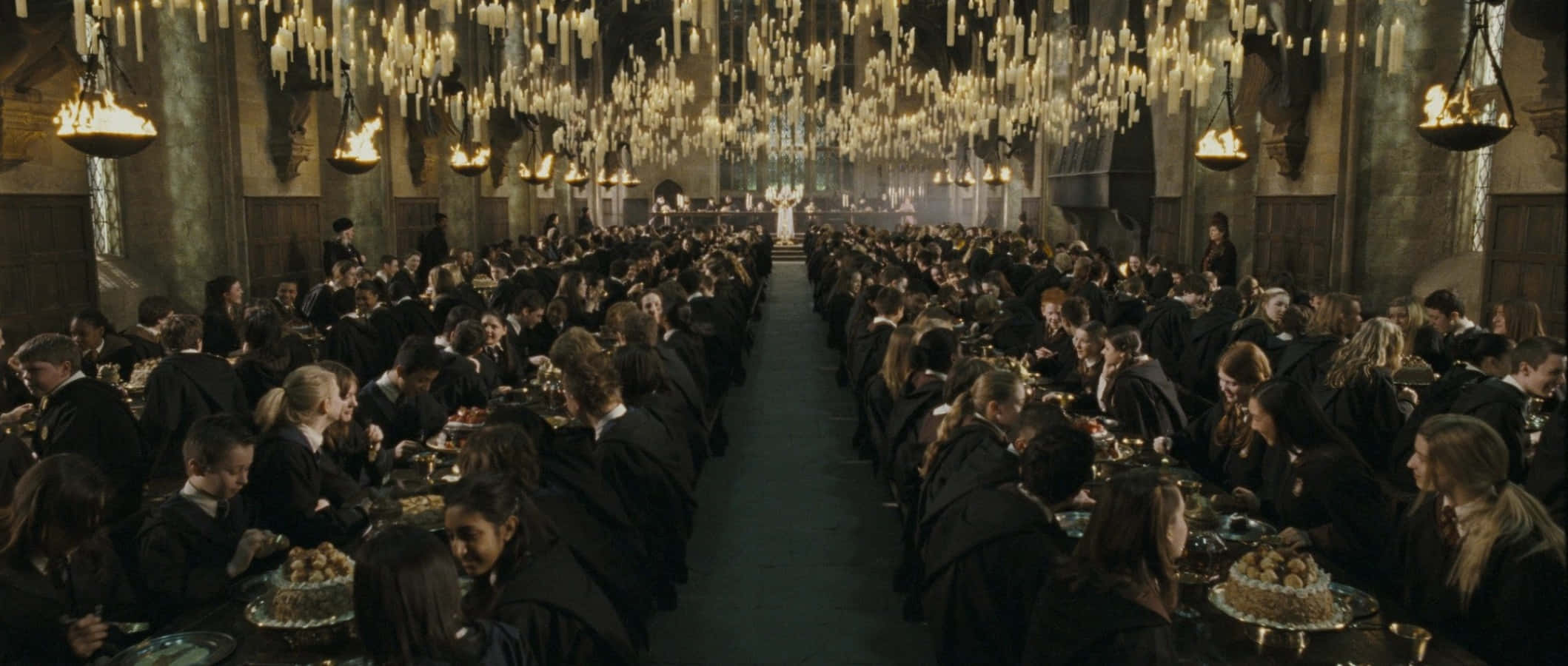 The Hogwarts Great Hall 桌布 壁紙 — 身穿黑色長袍的學生在宏偉的石廳中，在層疊的蠟燭下用餐，廳內擺滿長桌，充滿溫暖的金色光芒。