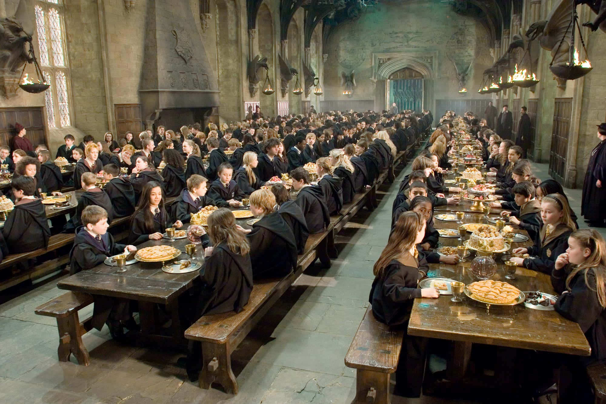 The Hogwarts Great Hall 桌布 壁紙 — 身穿黑色長袍的學生在高天花板和掛著火把的宏偉石廳中的長木桌上用餐。