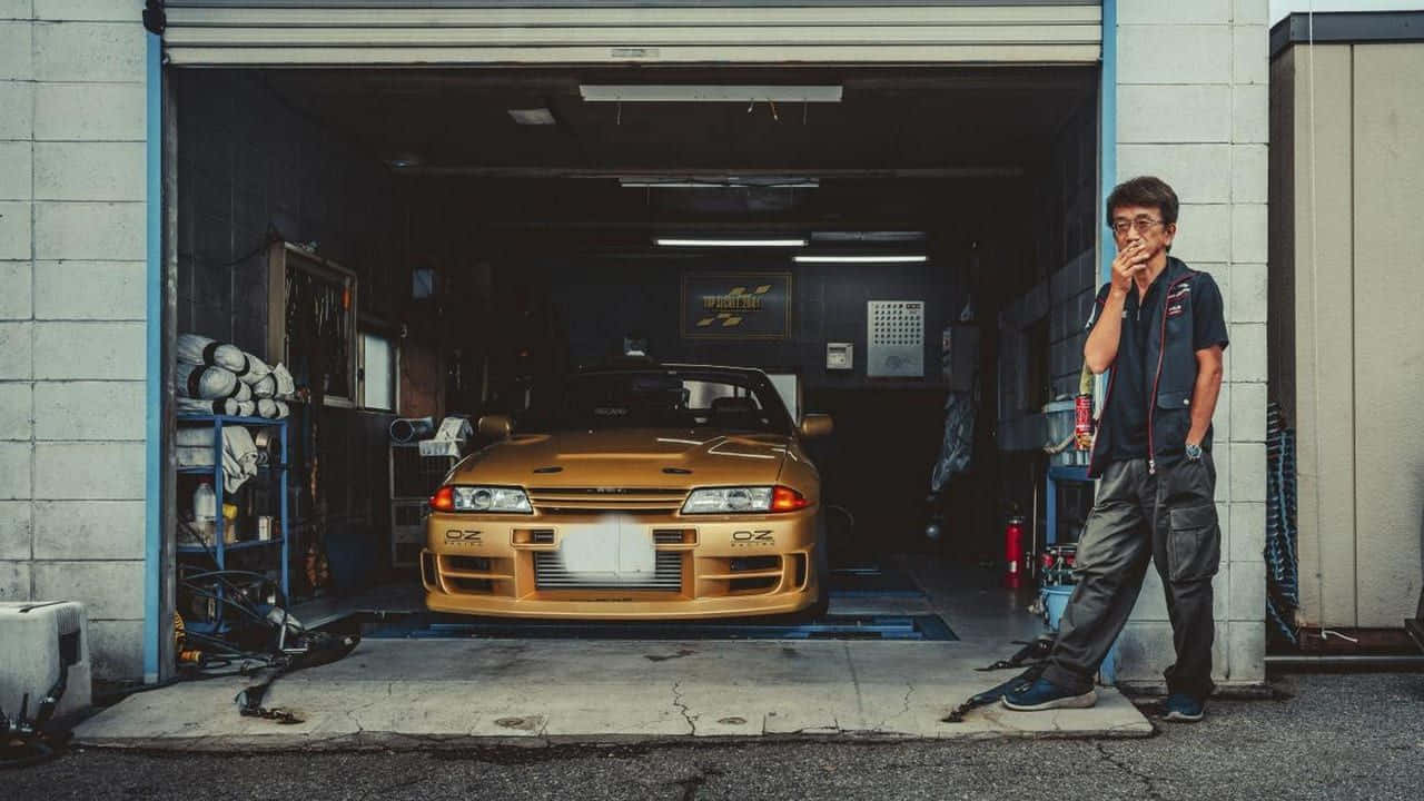 人像動態 桌布 壁紙 — 車庫裡的技師站在金色日產Skyline R32旁邊，周圍有工具和設備。