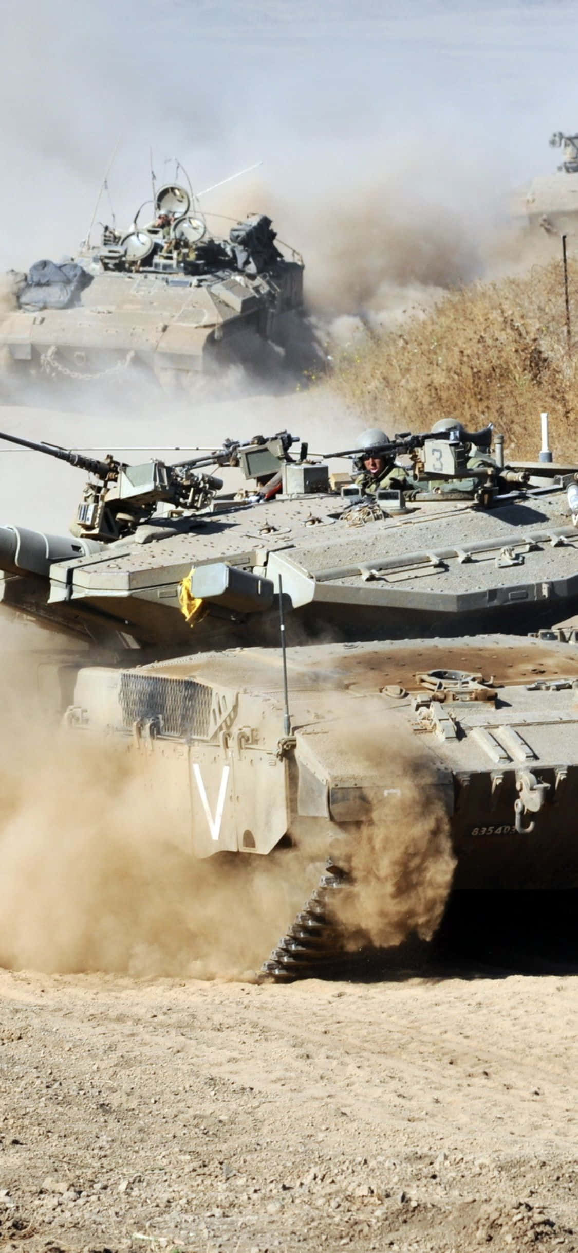 iPhone Xs Tanks 桌布 壁紙 — 軍用坦克在塵土飛揚的沙漠戰場上衝鋒，揚起沙塵和碎片雲。