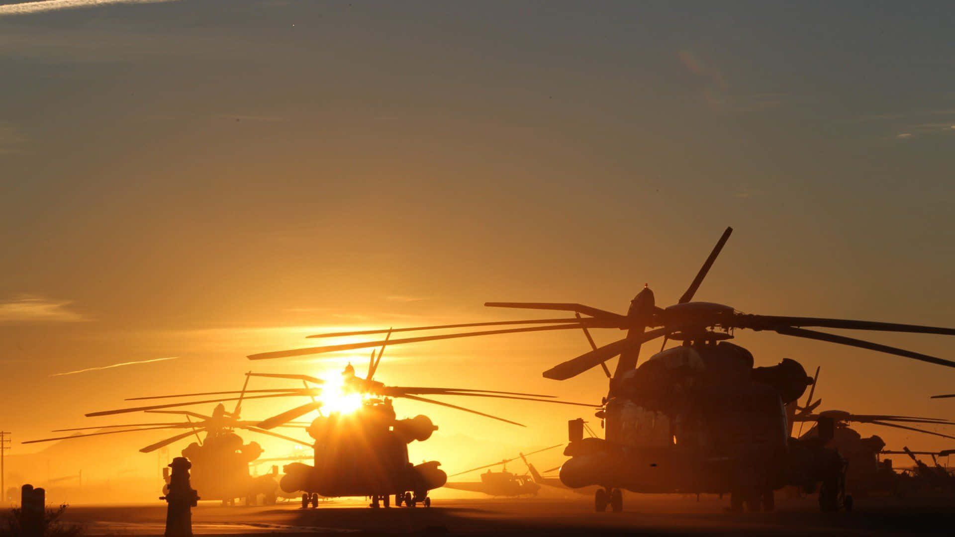 Helicopter 桌布 壁紙 — 軍用直升機的剪影靜止在停機坪上，夕陽在它們身後落下，散發出金色的光芒。