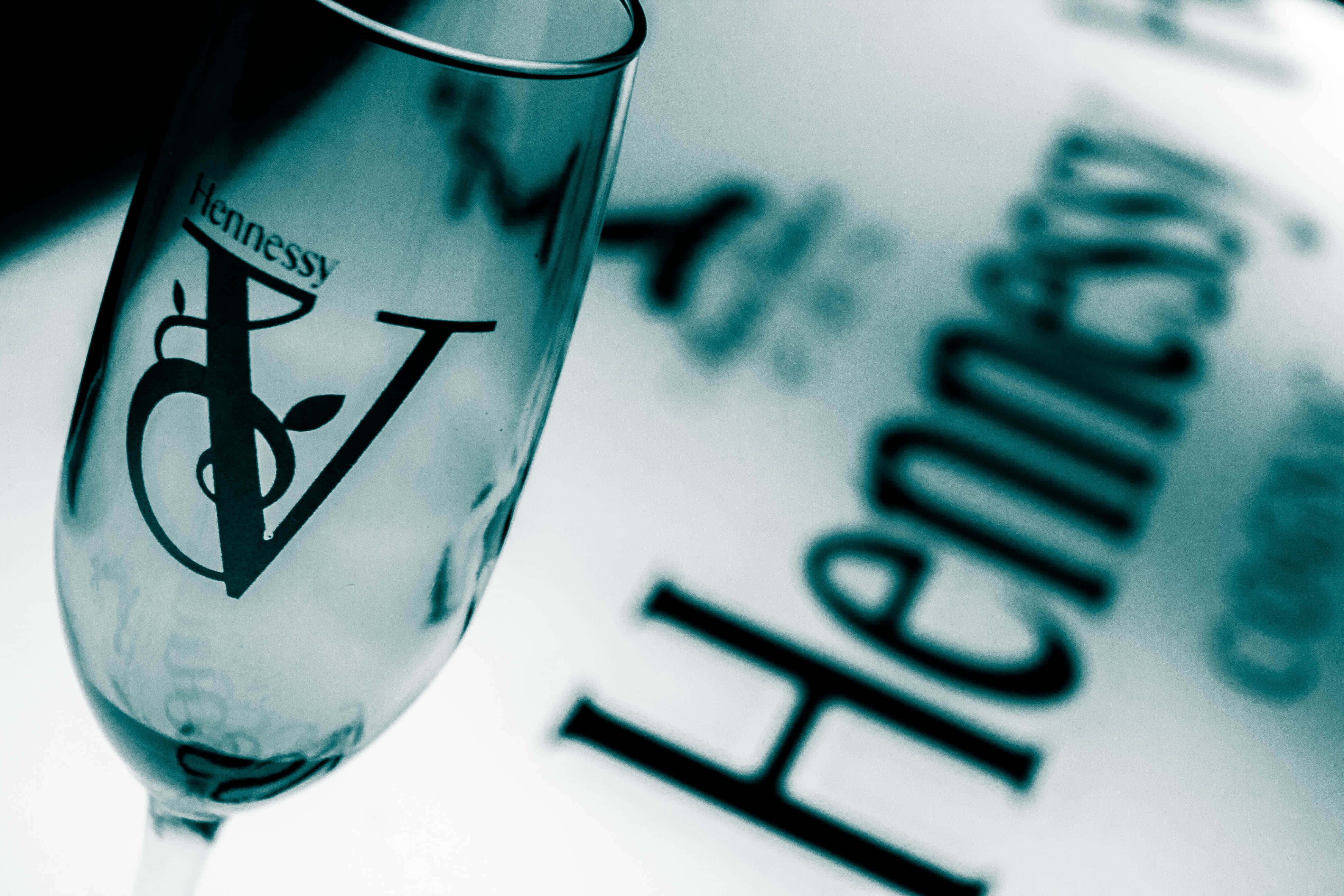 Hennessy Bottle 桌布 壁紙 — 軒尼詩品牌香檳杯的特寫，放在模糊的軒尼詩標誌表面上，帶有冷藍色調。