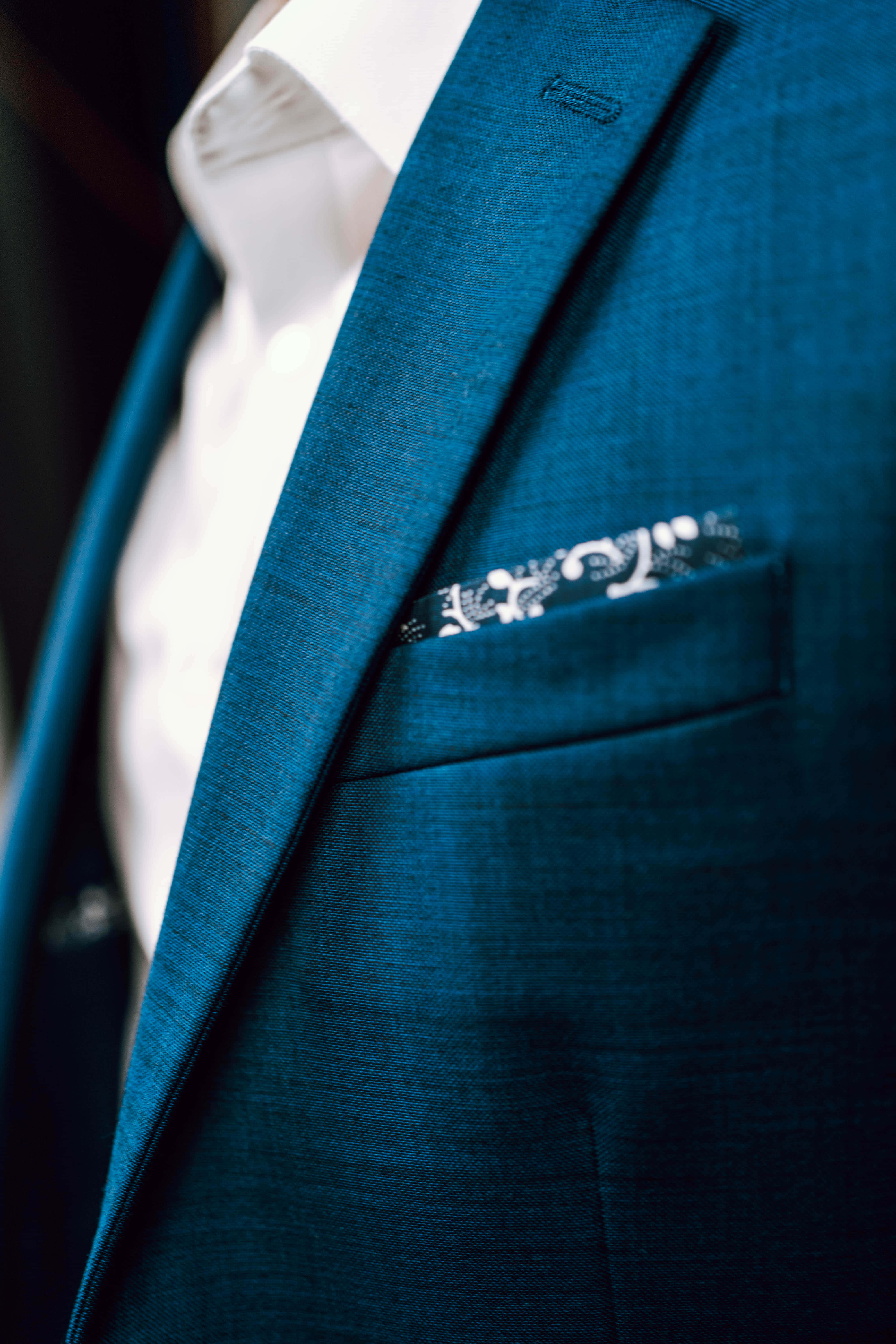 suit 桌布 壁紙 — 近距離拍攝一件帶有黑色花卉刺繡白色袋巾的藍綠色紋理西裝外套。