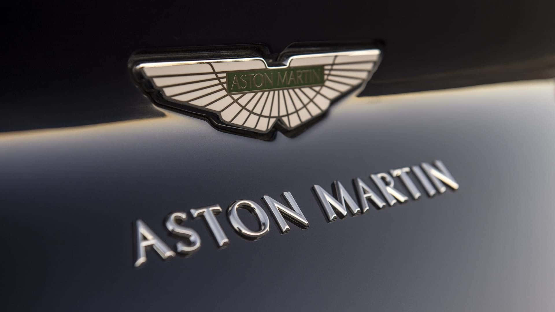 奧斯頓馬丁 桌布 壁紙 — 近距離拍攝一個光滑的Aston Martin徽章和字體，位於帶有金屬光澤的深色汽車引擎蓋上。