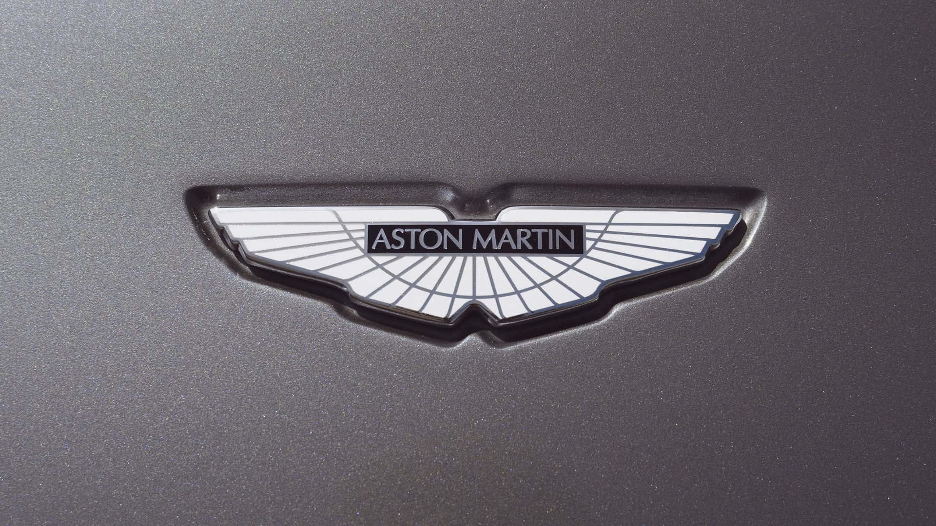 奧斯頓馬丁 桌布 壁紙 — 近距離拍攝一個光滑的銀色Aston Martin翼形標誌徽章，位於啞光深灰色汽車表面。