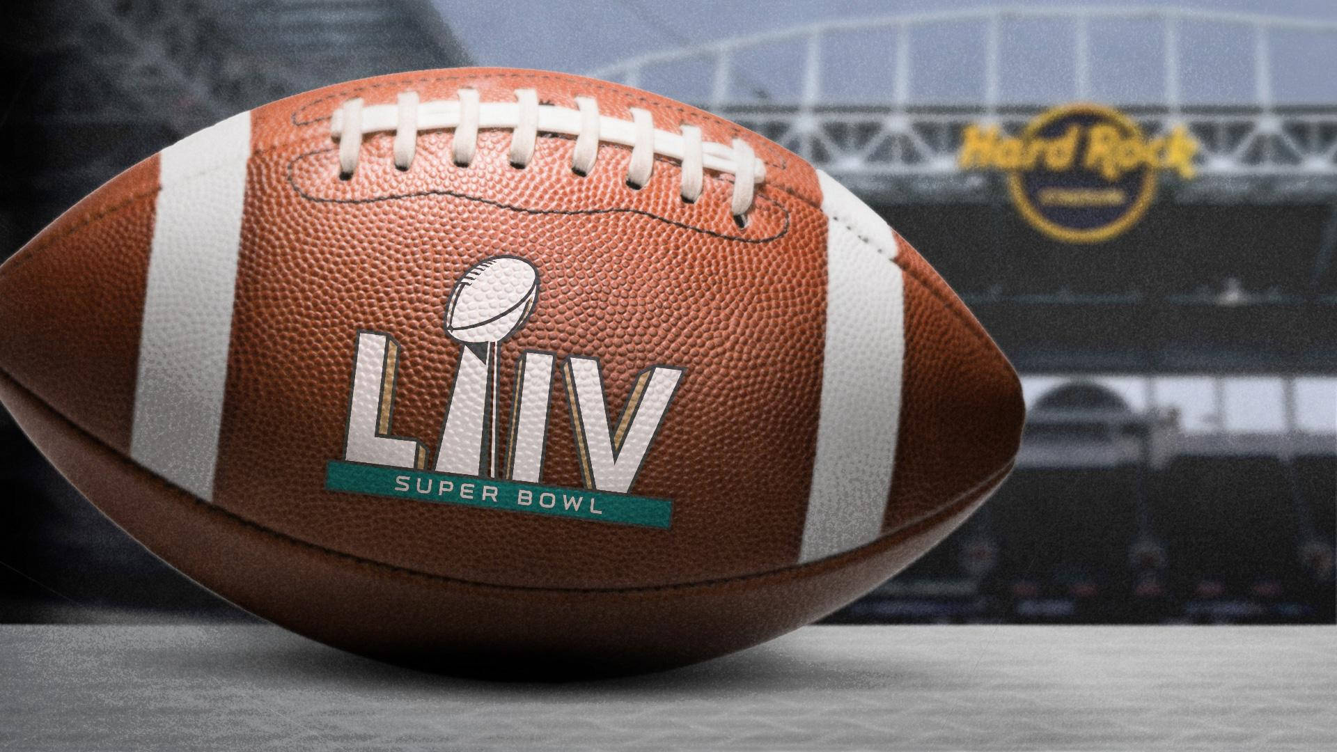 Super 桌布 壁紙 — 近距離拍攝一個棕色皮革 Super Bowl LIV 足球，帶有白色鞋帶和標誌，放置在灰色表面上，背景是模糊的體育場。