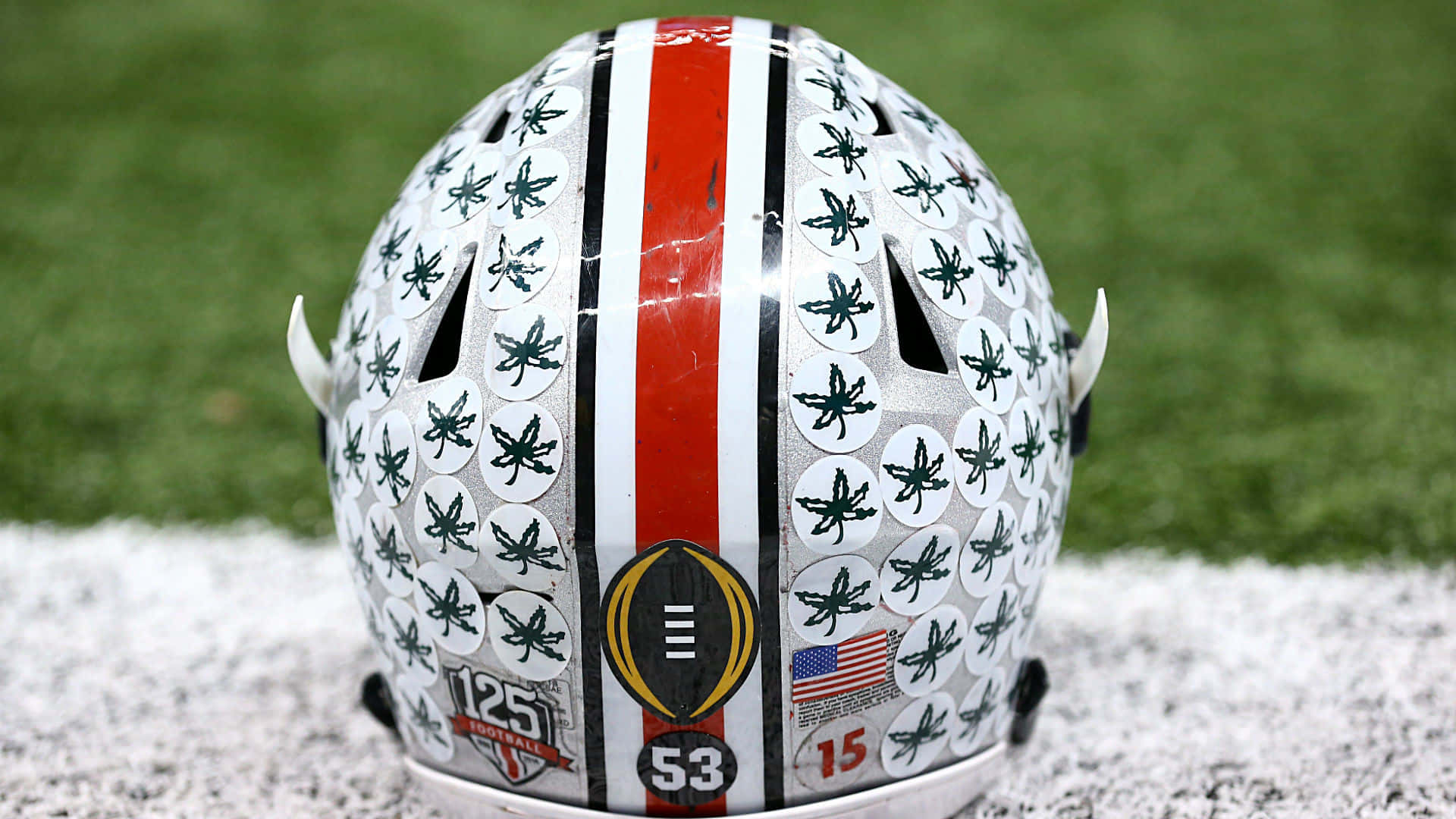 Cool Ohio State 桌布 壁紙 — 近距離拍攝一個美式足球頭盔，上面貼滿大麻葉貼紙，擱在積雪的草地上，背景是綠色草地。