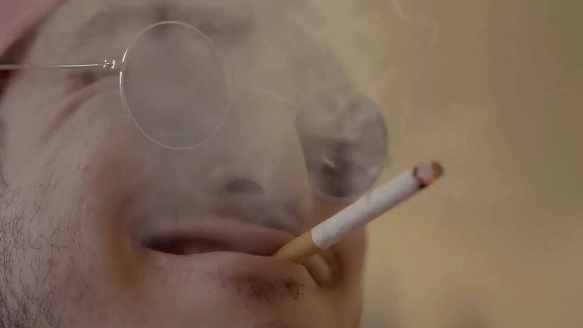 高清 桌布 壁紙 — 近距離拍攝一名戴圓框眼鏡的男子，嘴唇夾著點燃的香煙，呼出煙霧。