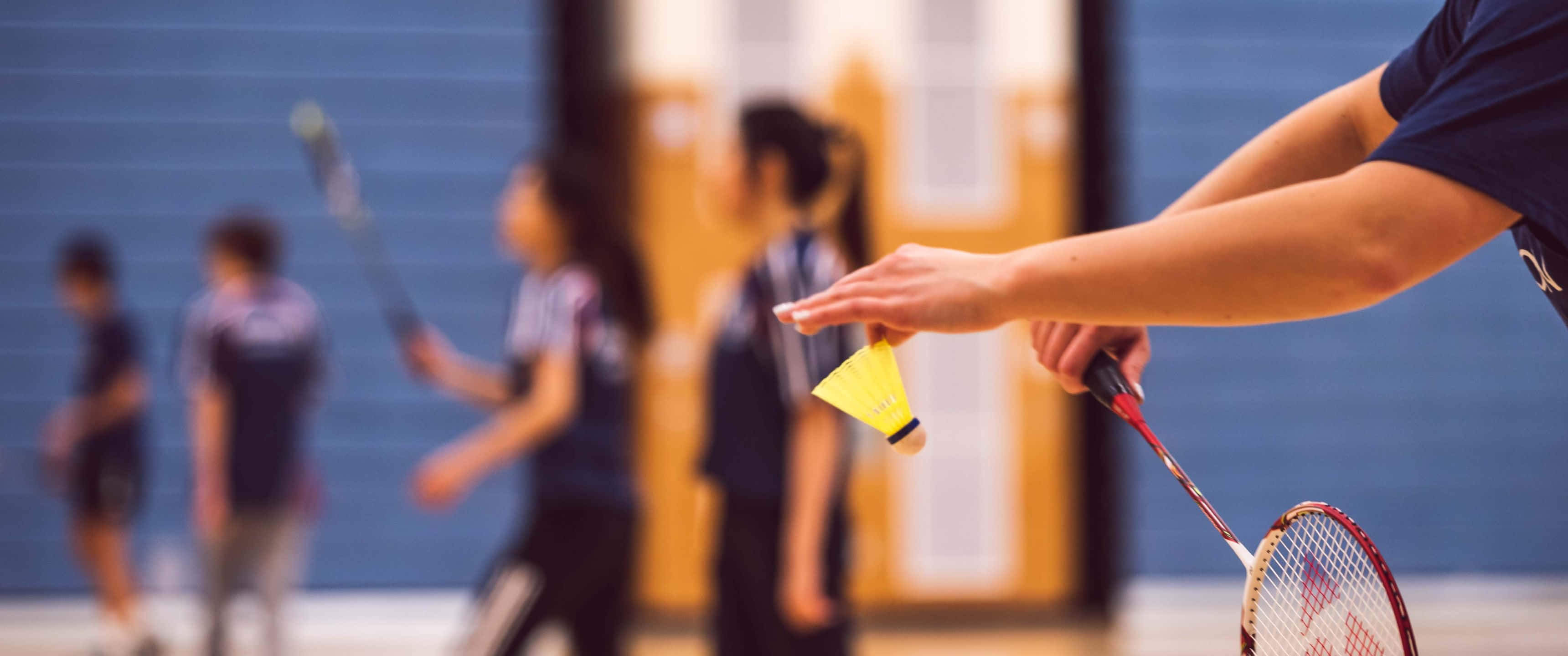 3440x1440p Badminton 桌布 壁紙 — 近距離拍攝一名球員手持羽毛球拍和羽毛球，背景中有隊友模糊的身影，在室內場地上。