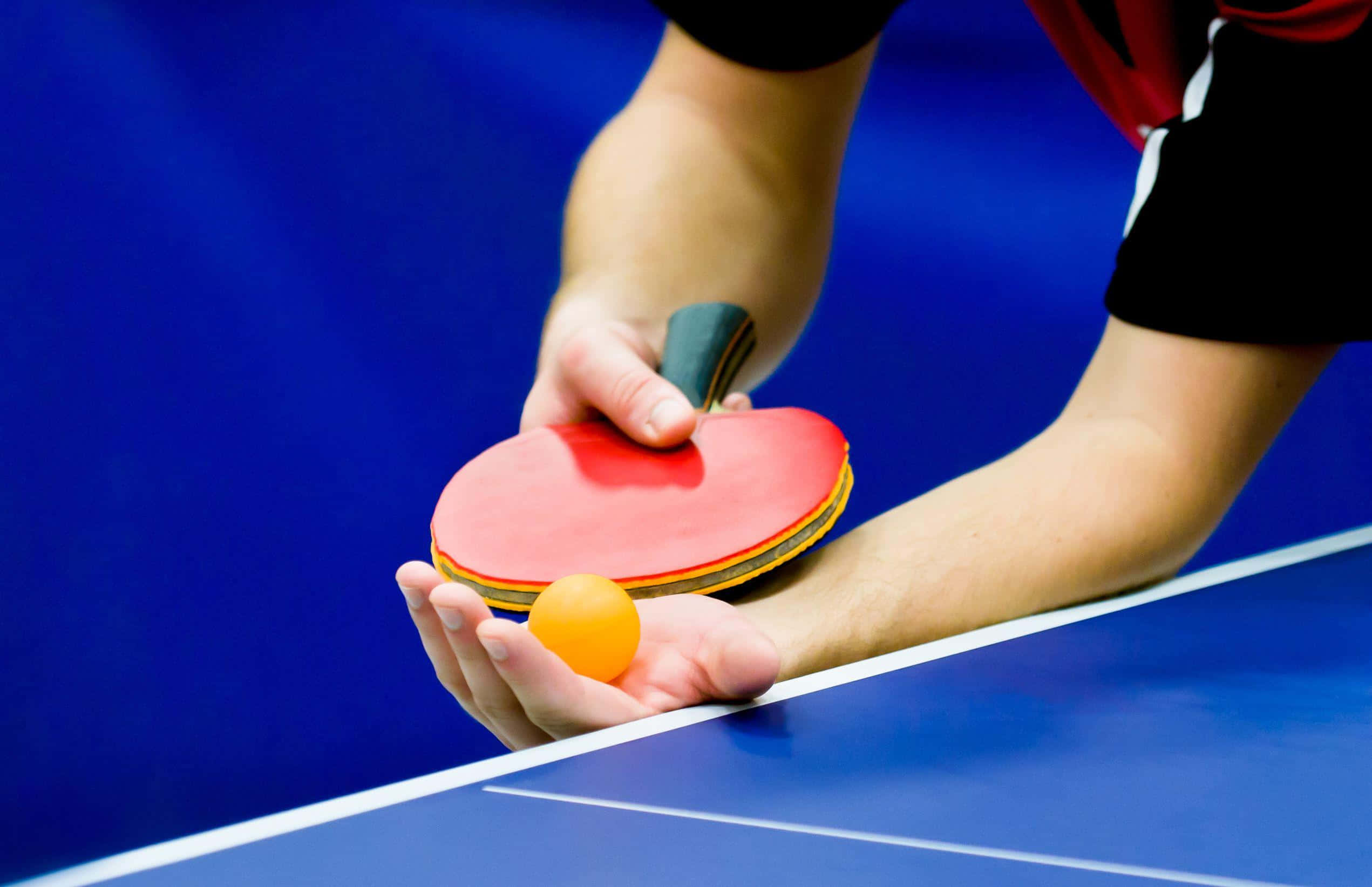 Table Tennis 桌布 壁紙 — 近距離拍攝一名球員的手，在帶有白色線條的藍色球桌上方握著紅色乒乓球拍和球。