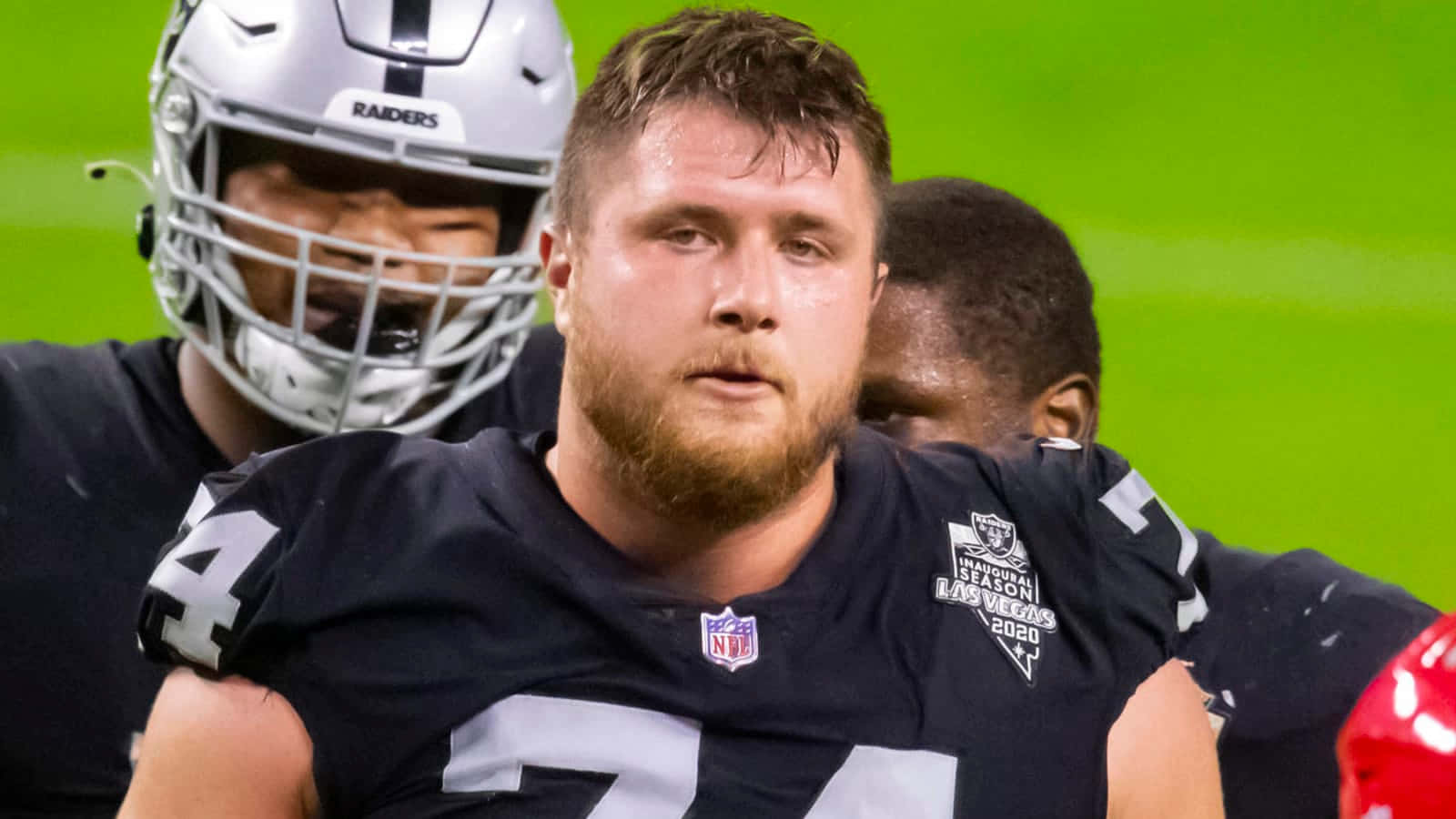 足球 桌布 壁紙 — 近距離拍攝一名穿著Raiders球衣的NFL美式足球員，背景有隊友，在明亮的綠色球場上。