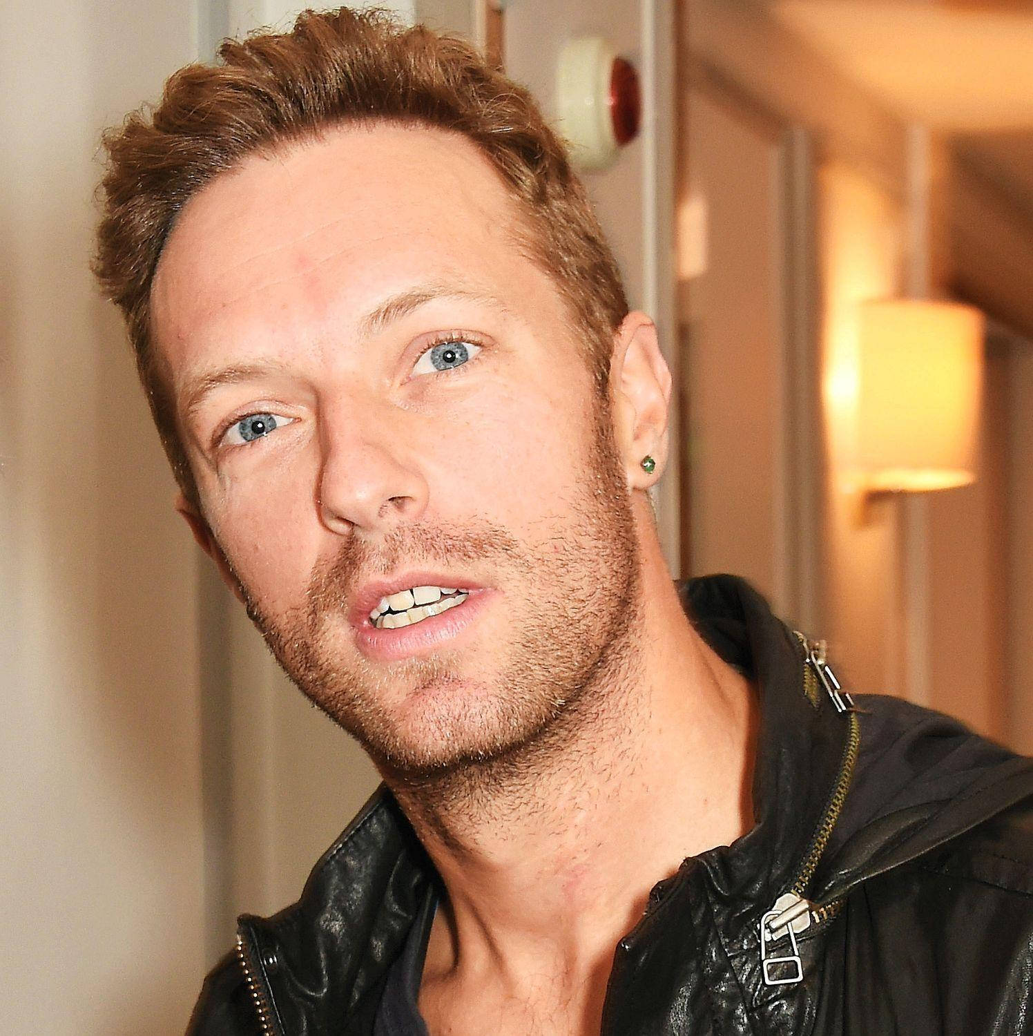 Chris Martin 桌布 壁紙 — 近距離拍攝一名藍眼睛、鬍渣未刮乾淨的男子，在室內溫暖燈光下穿著黑色皮夾克。