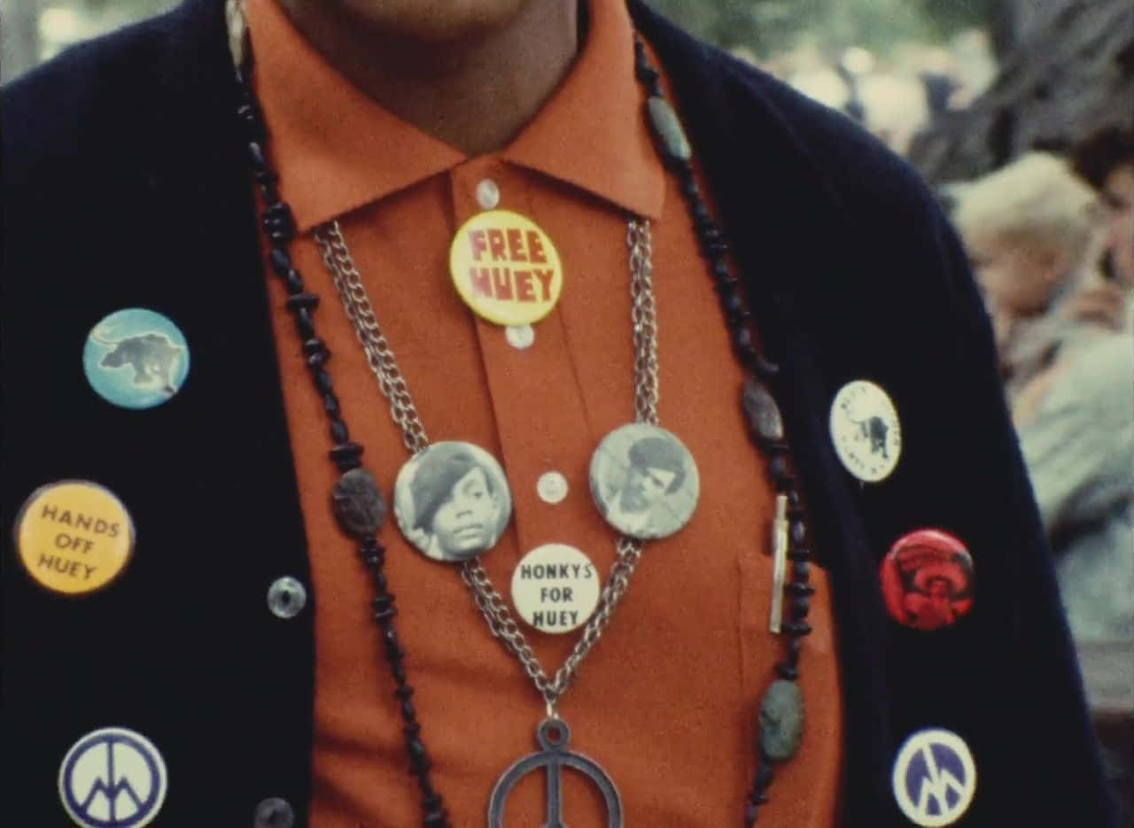 Black Panther Party 桌布 壁紙 — 近距離拍攝一名身穿橙色襯衫和黑色開襟衫的人，衣服上佈滿抗議徽章和珠鏈。