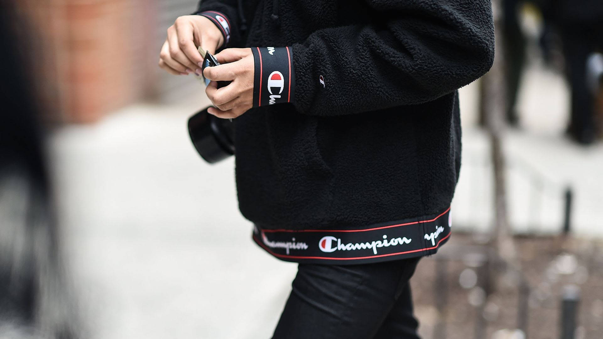 Champion 桌布 壁紙 — 近距離拍攝一名身穿黑色Champion刷毛外套的人，在行人道上調整相機帶。
