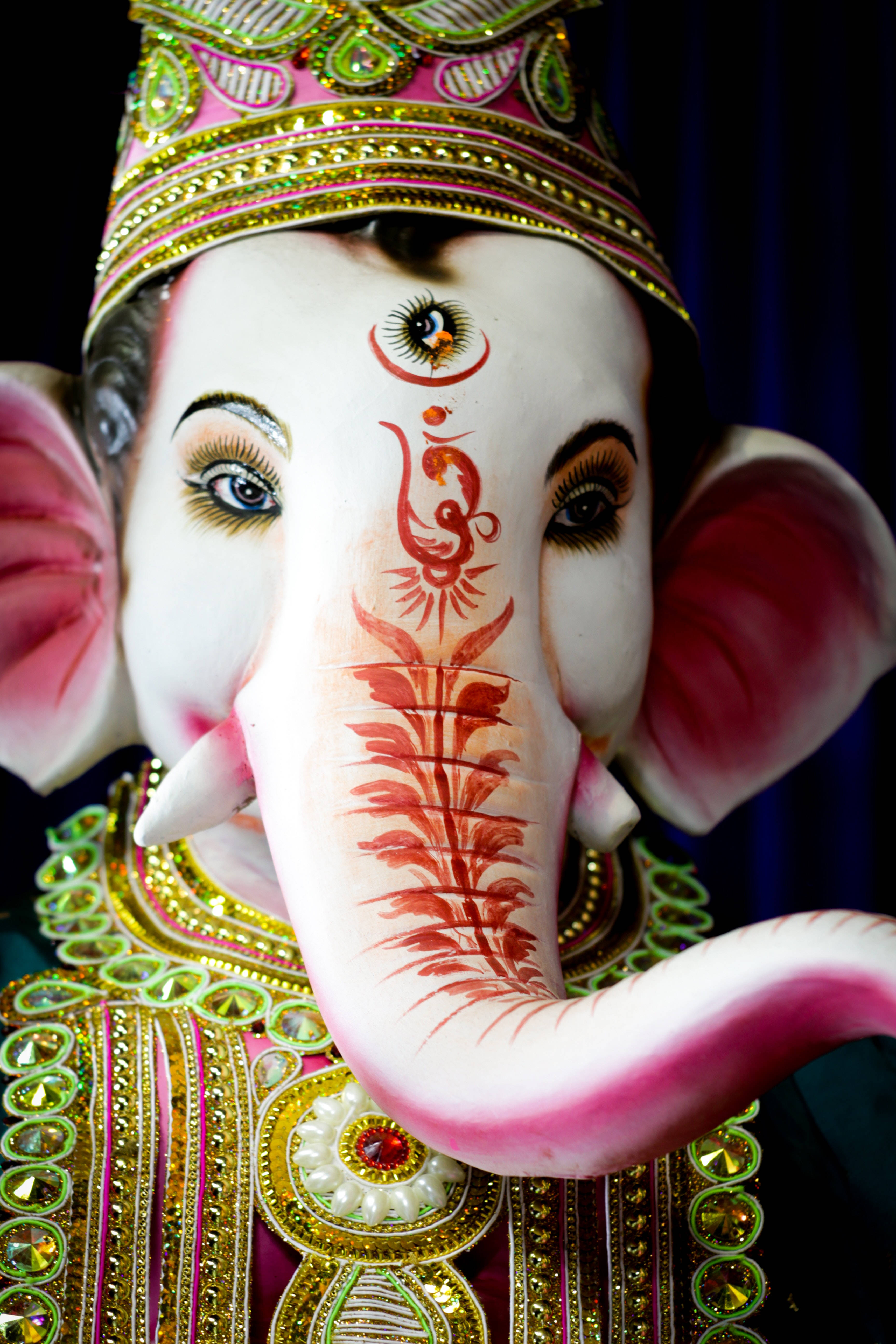 ganesh 4K 桌布 壁紙 — 近距離拍攝一尊色彩繽紛的象神雕像，帶有粉紅色耳朵、金色皇冠和紅色指甲花圖案在象鼻上。