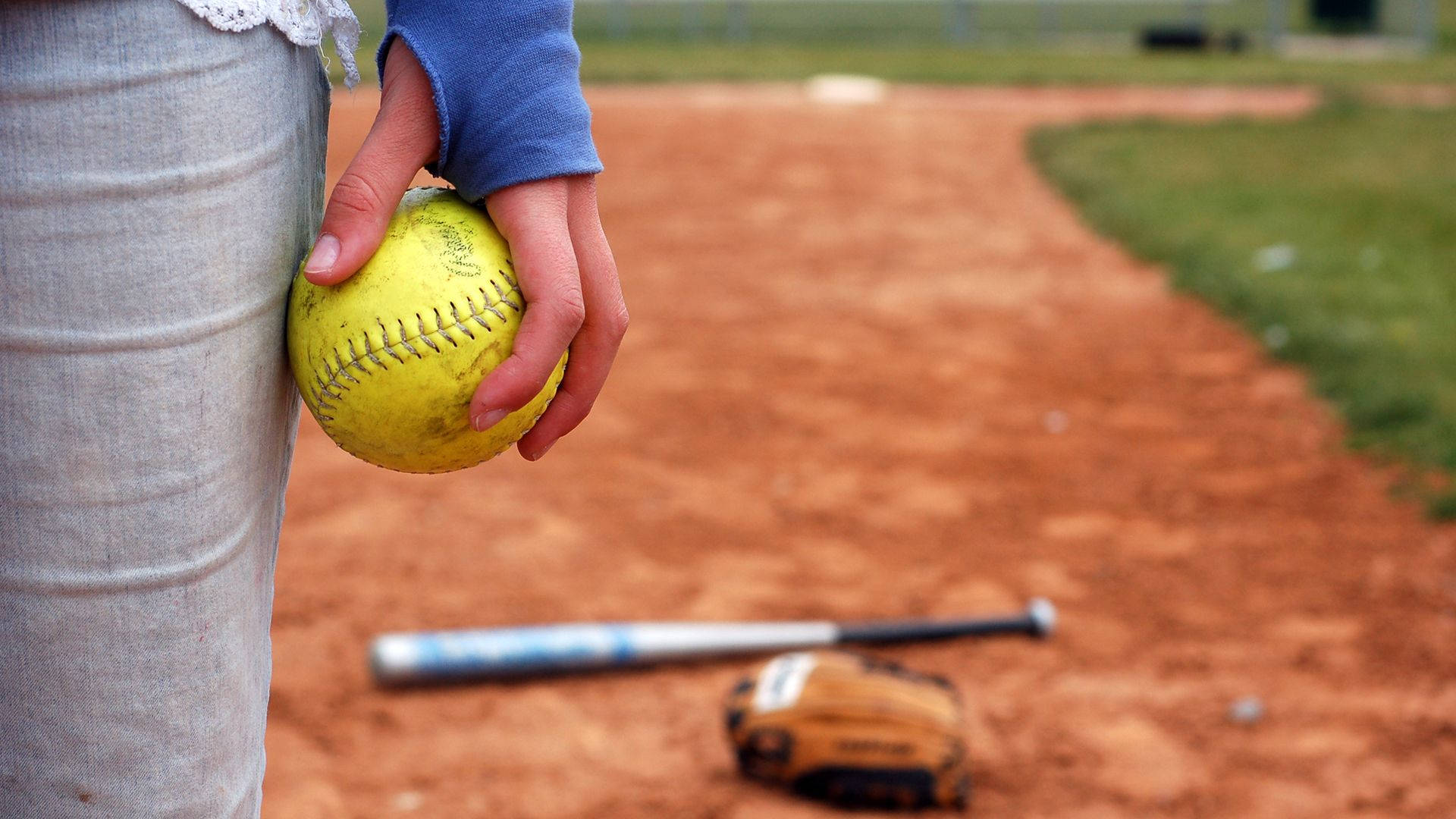 Softball 桌布 壁紙 — 近距離拍攝一隻手在泥濘的棒球場上握住一個黃色壘球，附近有球棒和手套。