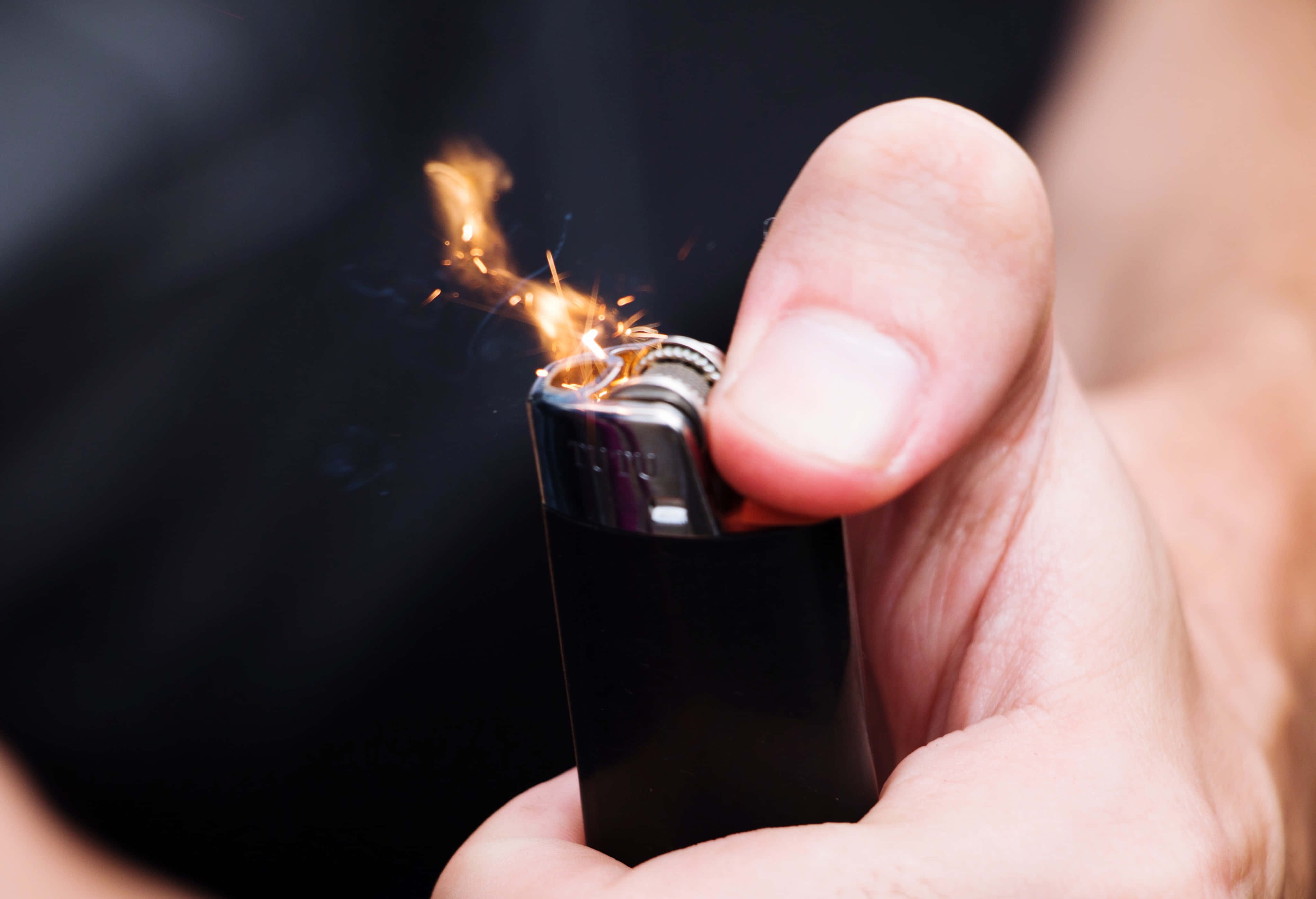 lighter 桌布 壁紙 — 近距離拍攝一隻手在黑暗背景下點燃一個黑色打火機，帶有明亮的橙色火花和火焰。