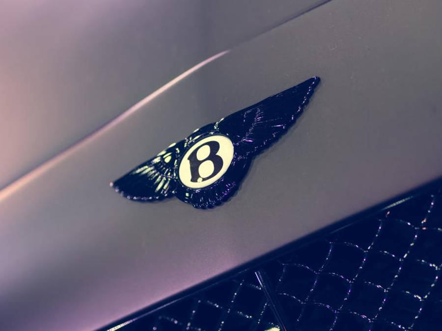 bentley HD 桌布 壁紙 — 近距離拍攝光澤的賓利帶翼 'B' 徽章，位於時尚金屬引擎蓋上，配有絎縫皮革飾邊。
