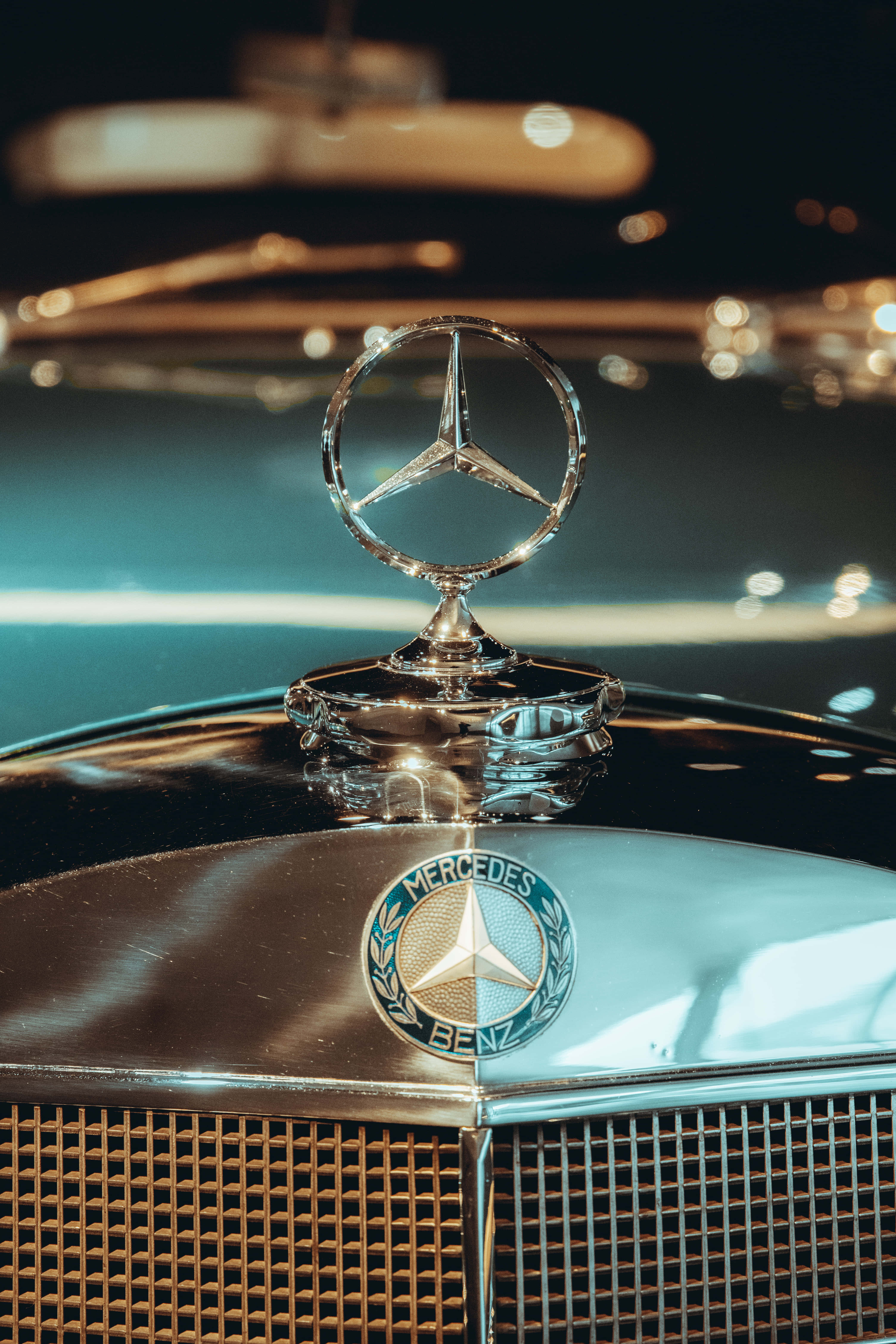 iPhone X Mercedes 桌布 壁紙 — 近距離拍攝在溫暖夜燈下閃閃發光的賓士引擎蓋標誌和格柵，背景呈散景效果。