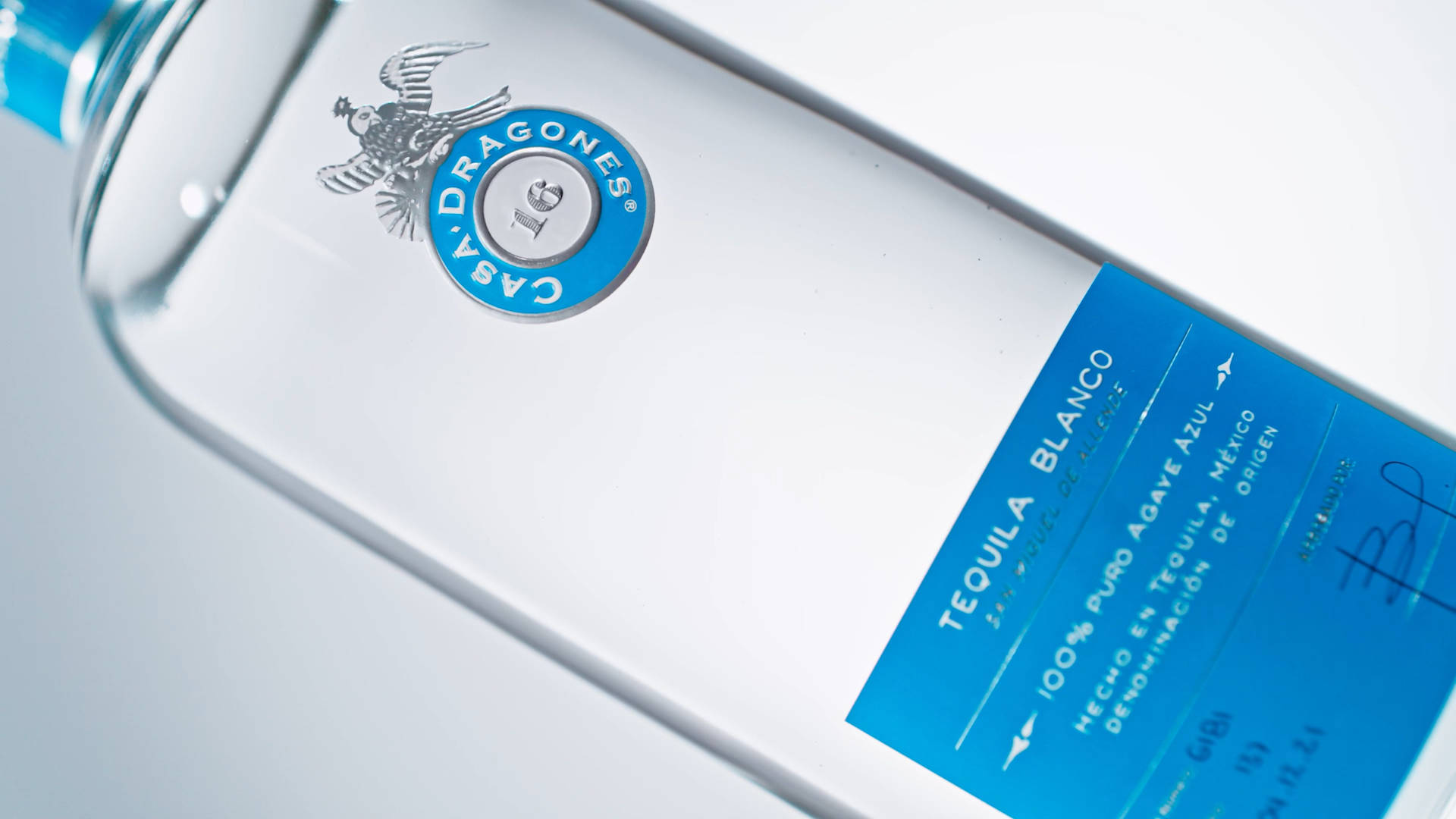 bottle 桌布 壁紙 — 近距離拍攝時尚的Casa Dragones龍舌蘭酒瓶，展示其藍色標籤和銀色鷹徽，背景為簡約白色。
