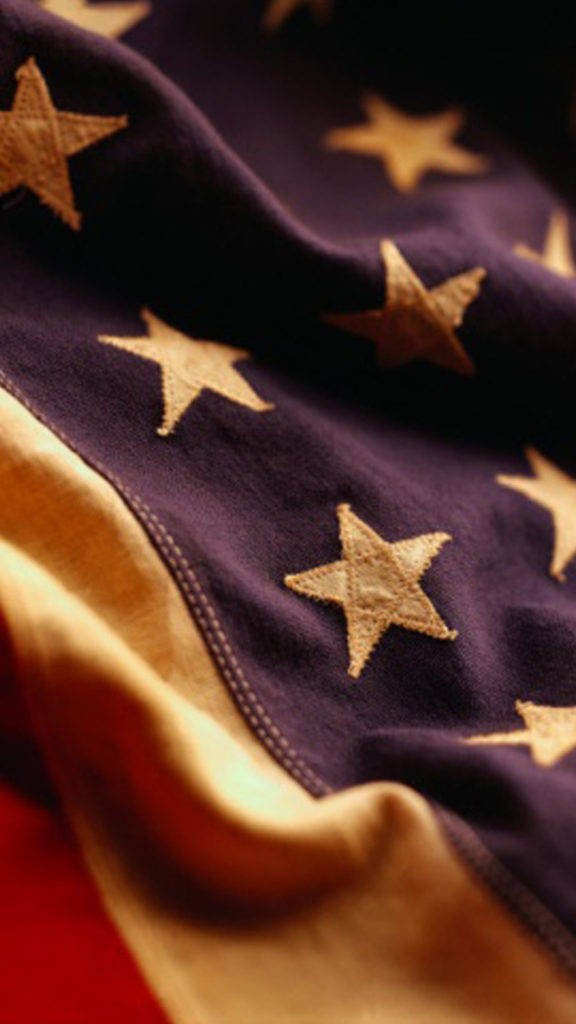 American Flag iPhone 桌布 壁紙 — 近距離拍攝有紋理的美國國旗，深藍色布料上有金色星星，紅色條紋在背景中柔和模糊。