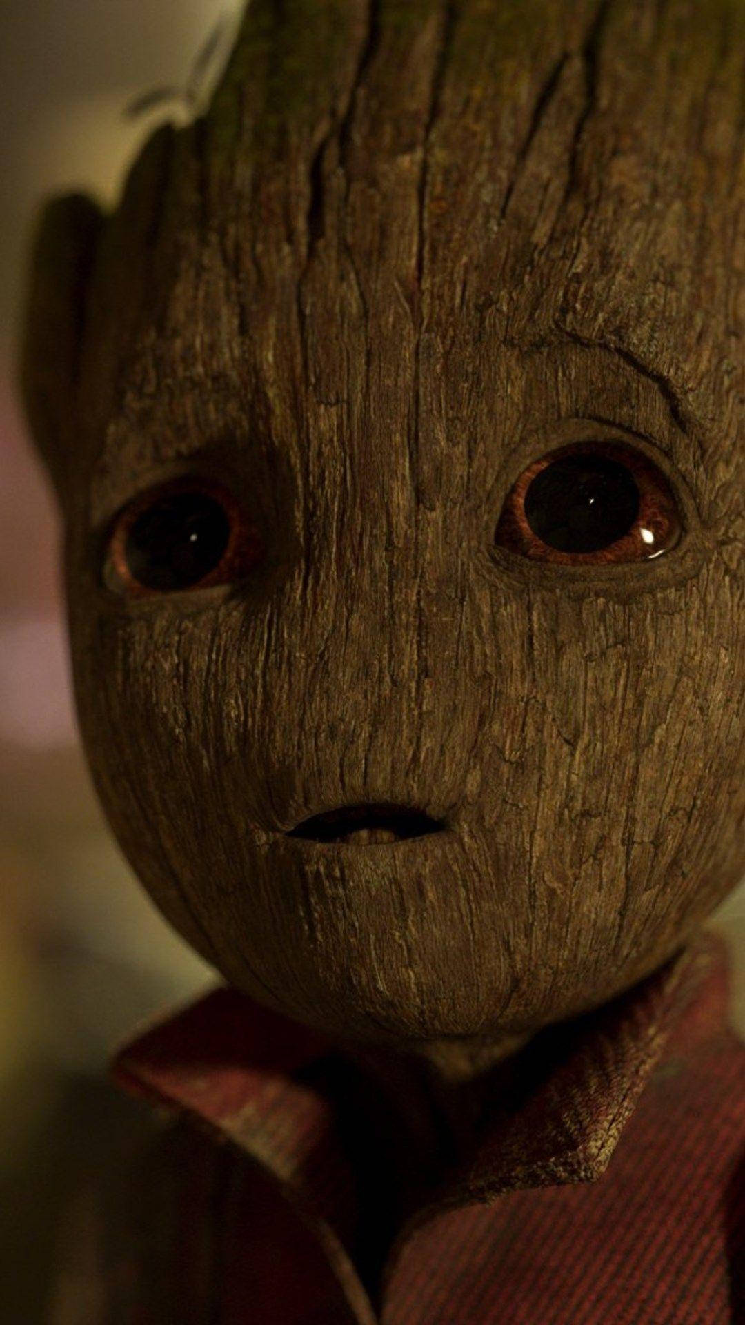 Baby Groot 桌布 壁紙 — 近距離拍攝格魯特紋理木質臉部，有大黑眼睛和紅色有領襯衫，處於溫暖昏暗燈光下。
