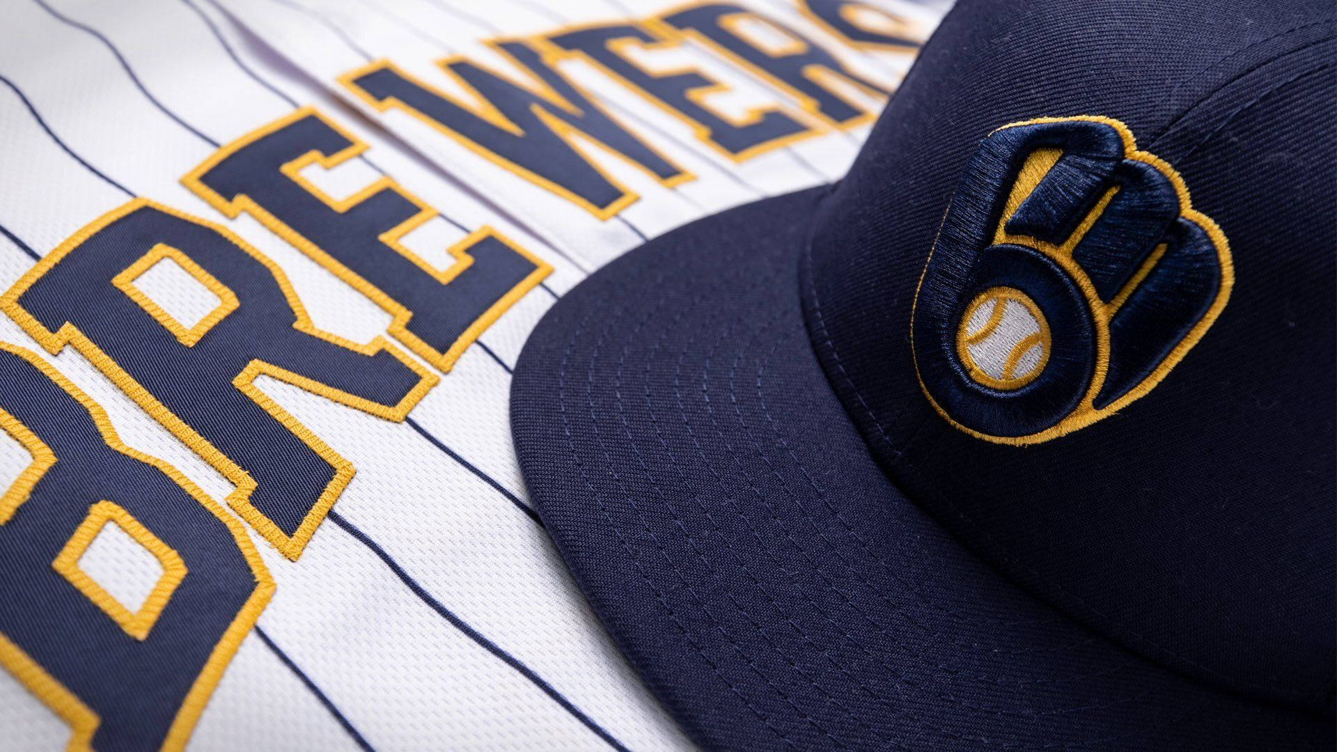 Brewers 桌布 壁紙 — 近距離拍攝海軍藍色密爾瓦基釀酒人棒球帽，放在白色細條紋球衣上，帶有粗體海軍藍色和金色字樣。