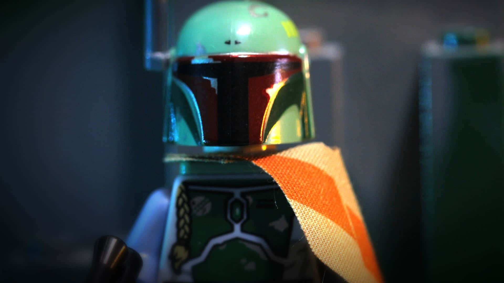 Jango Fett 桌布 壁紙 — 近距離拍攝的LEGO波巴·費特人偶，配備反光頭盔和橙色圍巾，背景為暗色模糊效果。