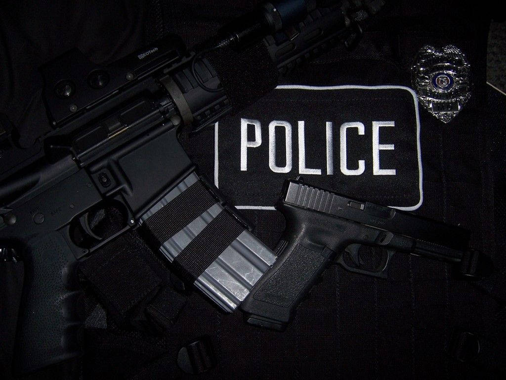 警察 桌布 壁紙 — 近距離拍攝的警察裝備，包括步槍手槍和徽章，放在帶有「POLICE」補丁的黑色制服上。