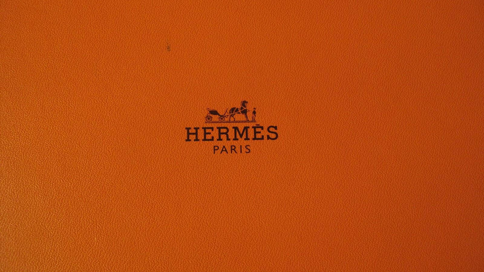 Hermes 桌布 壁紙 — 近距離拍攝的豐富橙色紋理皮革表面，帶有黑色 Hermès Paris 標誌和馬車圖案徽章。