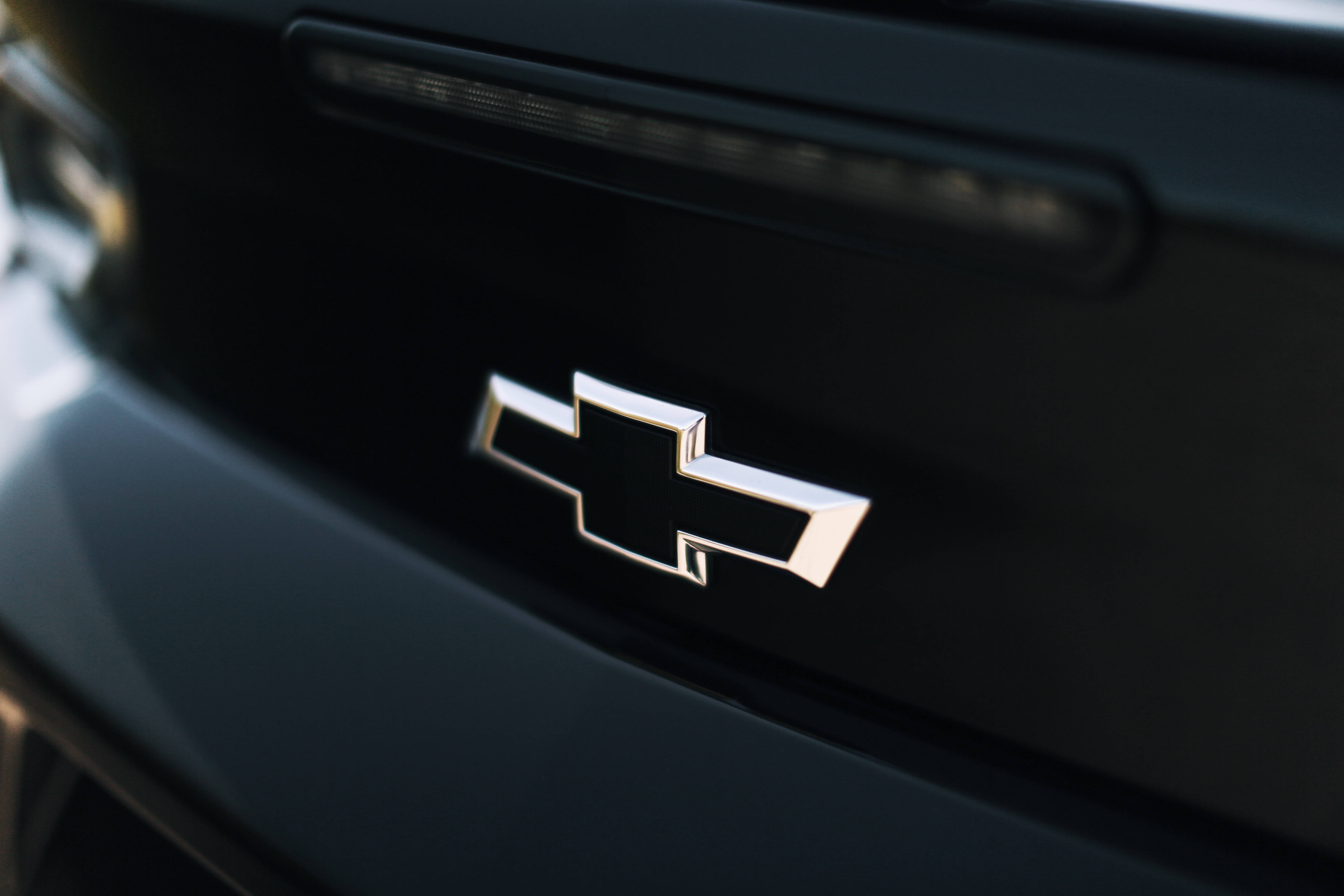 Chevrolet Logo 桌布 壁紙 — 近距離拍攝閃亮鍍鉻雪佛蘭蝴蝶結標誌，位於時尚黑色汽車後車廂上，柔和陽光突顯其金屬邊緣。