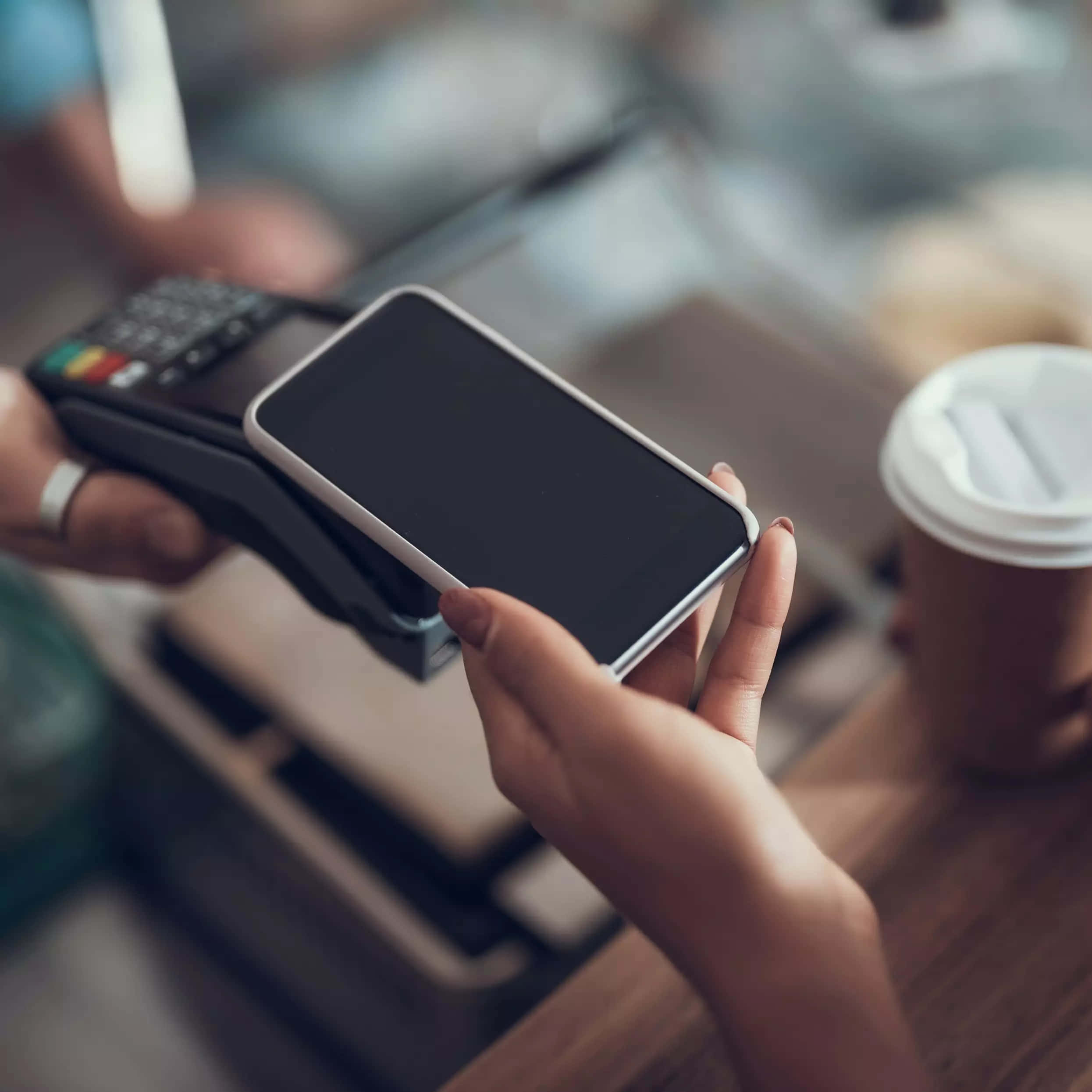 iPhone 桌布 壁紙 — 近距離拍攝雙手在咖啡店櫃檯的支付終端上輕觸智慧型手機，旁邊有一杯咖啡。