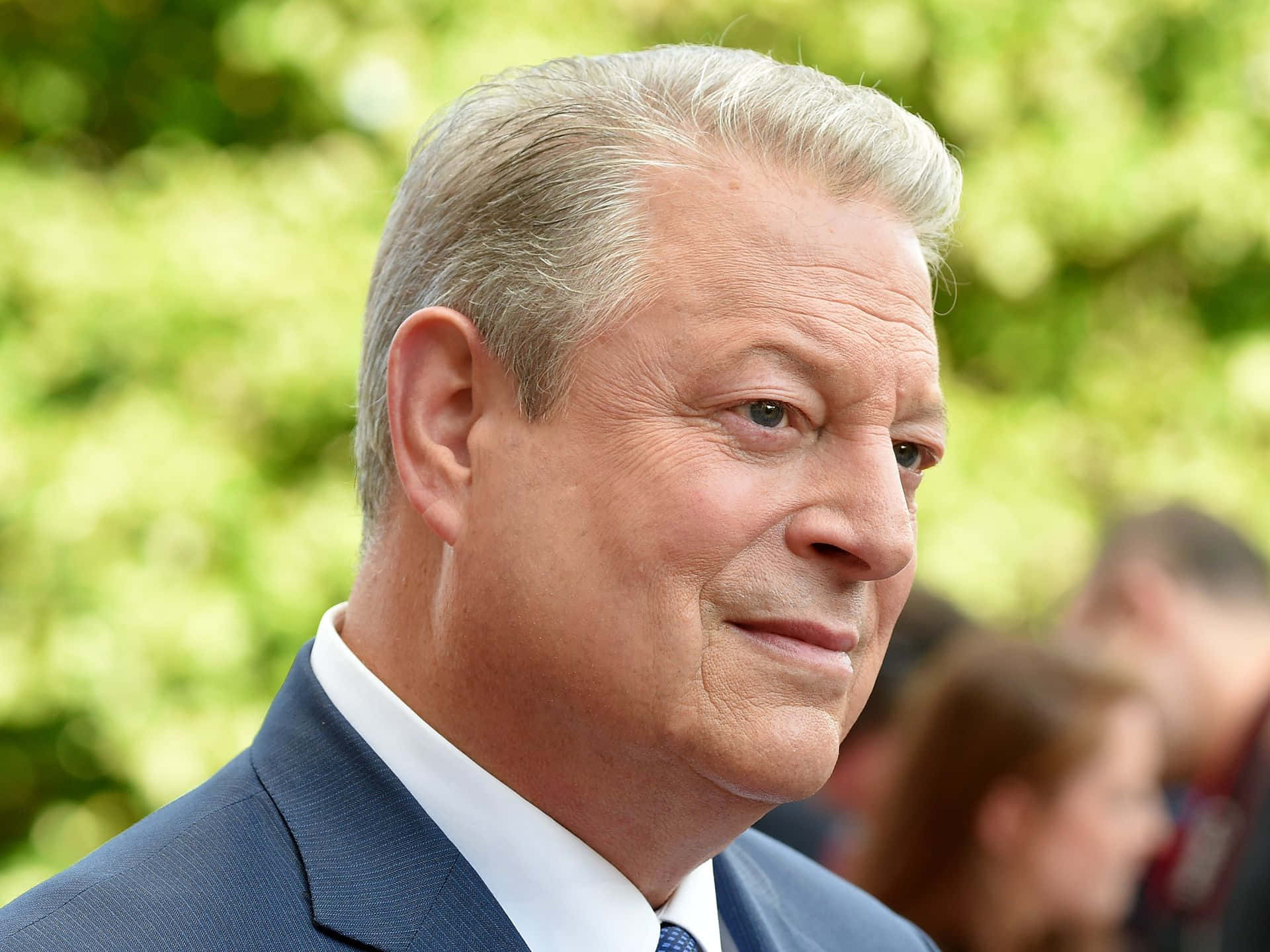 Celebrity 桌布 壁紙 — 近距離肖像，一位灰髮男子身穿藍色西裝，背景是模糊的綠色樹葉。