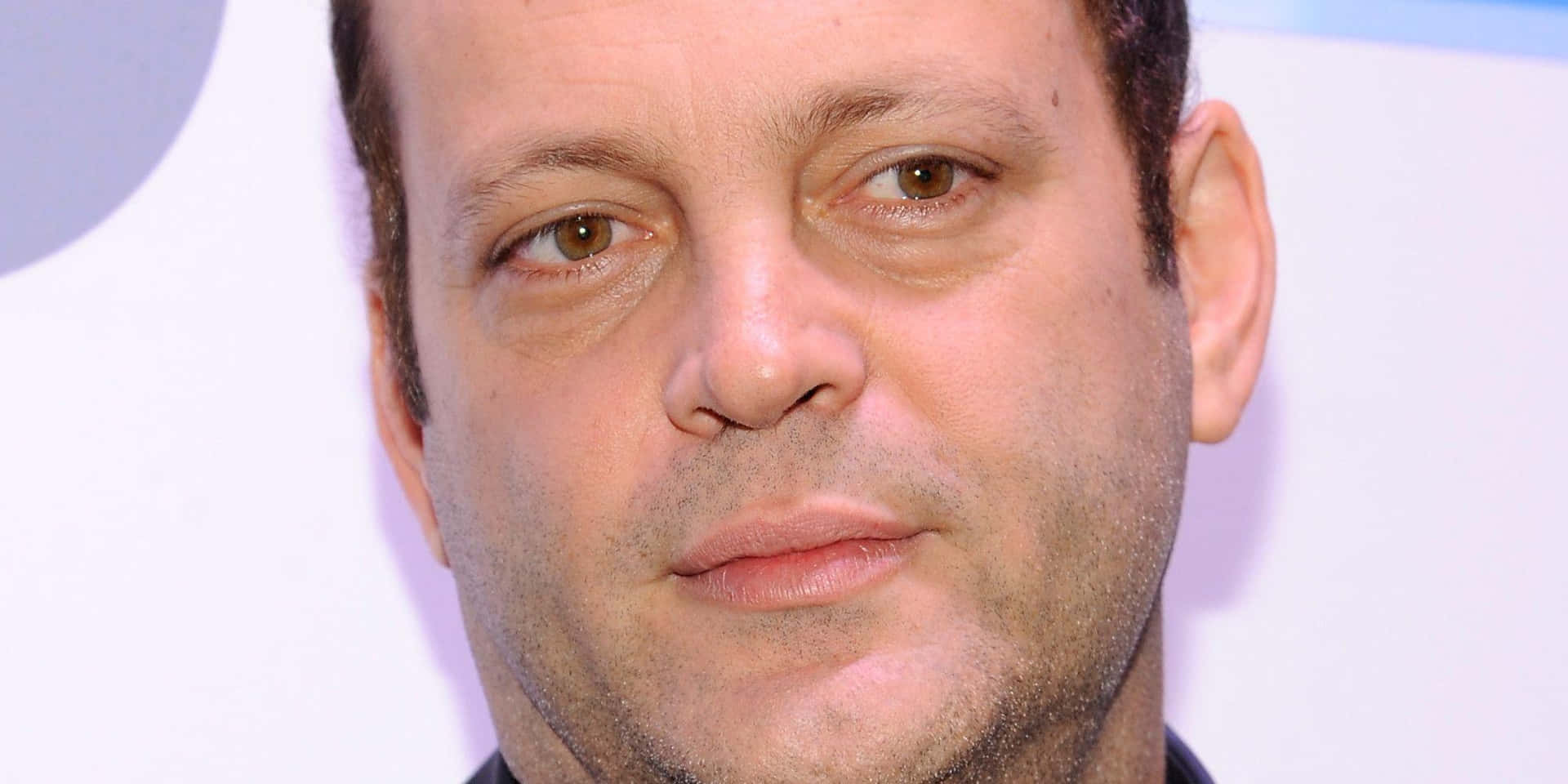Vince Vaughn 桌布 壁紙 — 近距離肖像，一名擁有棕色眼睛和鬍渣的男子，背景為柔和的白色和紫色。