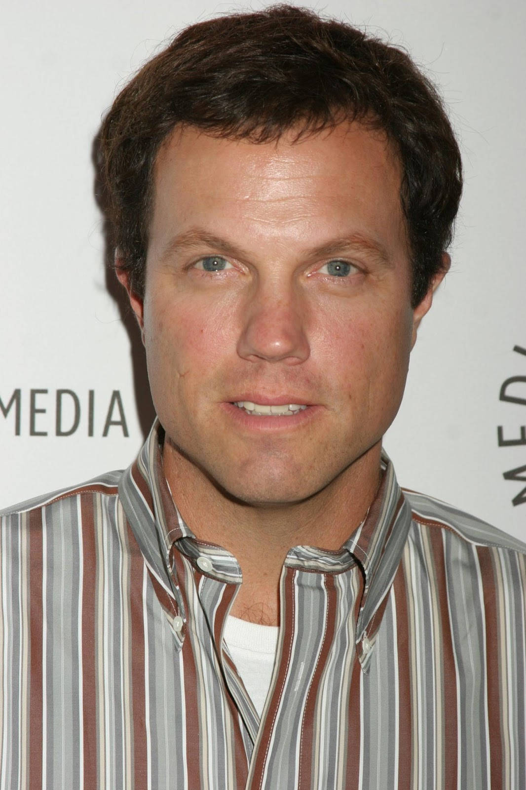 adam baldwin 桌布 壁紙 — 近距離肖像，一名棕色頭髮藍眼睛的男子穿著條紋襯衫，背景為白色並帶有部分文字。